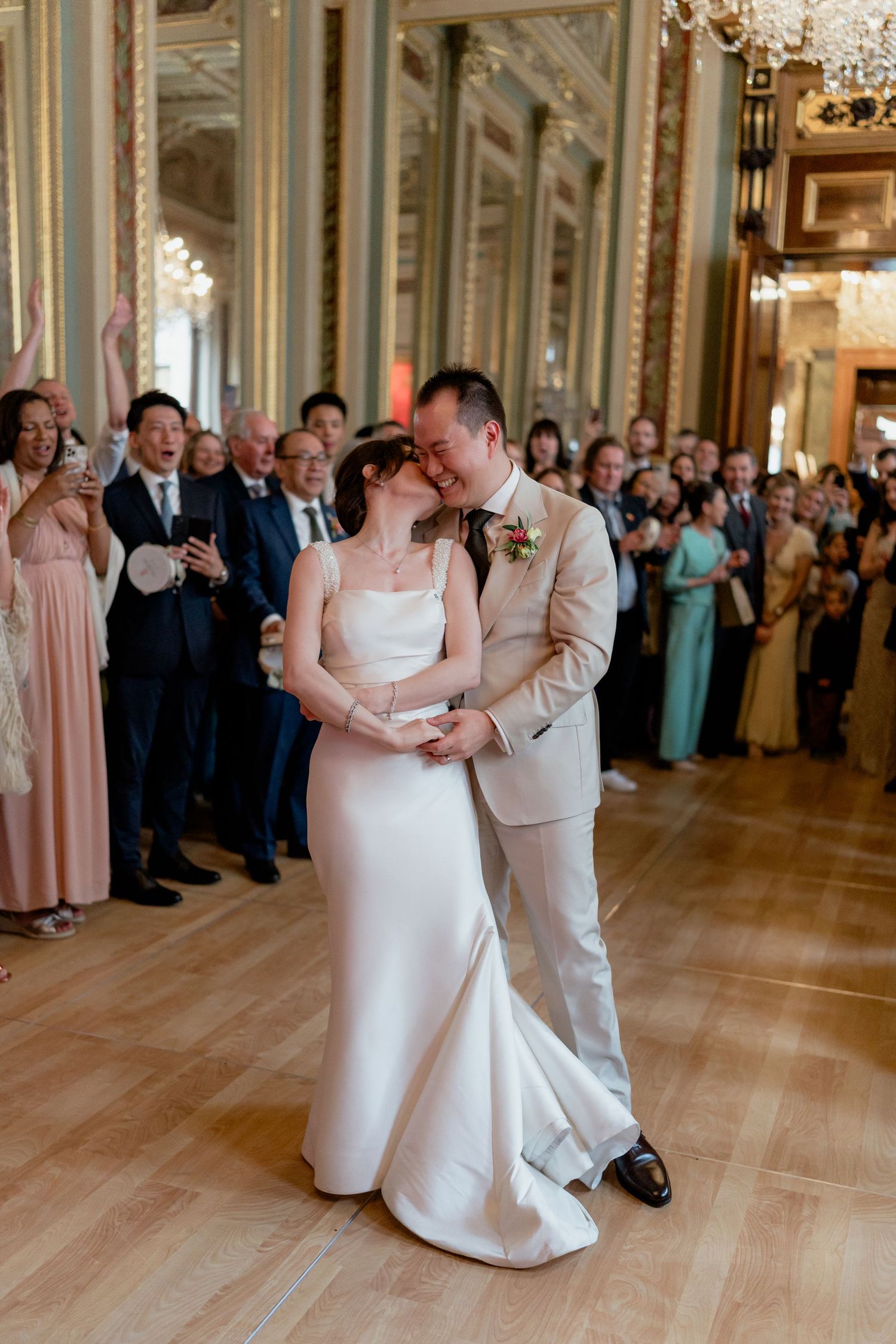 Bride and groom sharing their first dance in an ornate ballroom surrounded by cheering wedding guests.