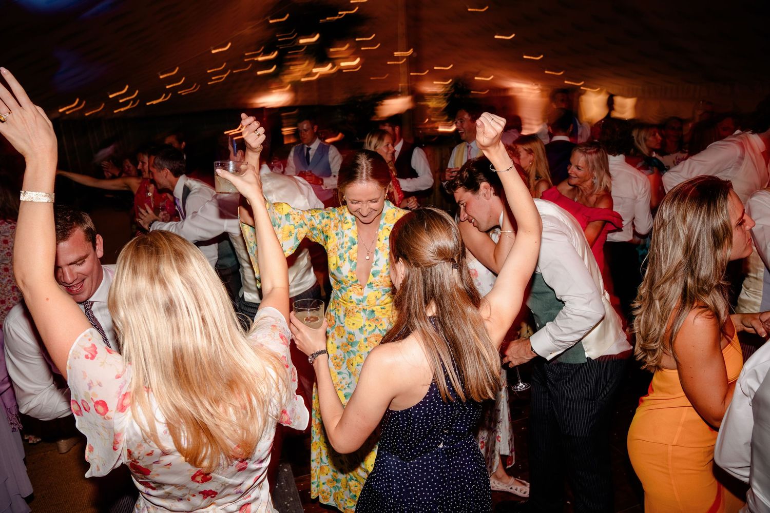 Energetic wedding guests dance together under warm lights on a crowded indoor reception dance floor.