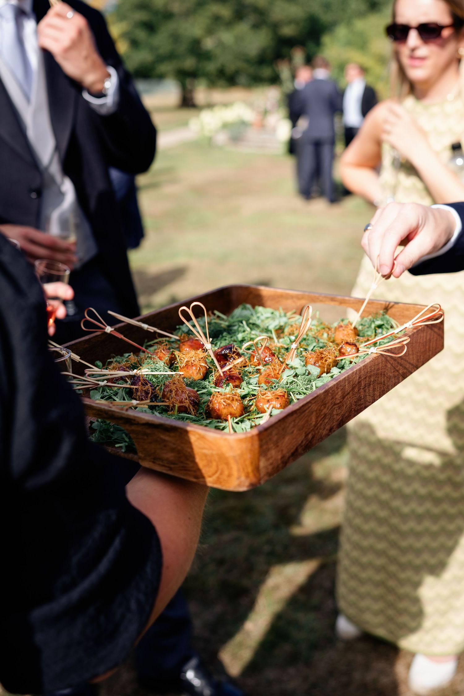 Guest reaches for fried canapé skewer from rustic wooden serving tray at outdoor wedding reception.