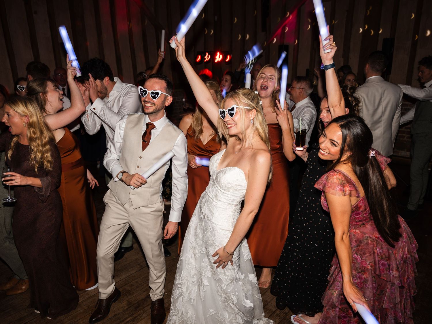 Bride and groom dance joyfully with guests waving glow sticks at a lively wedding reception.