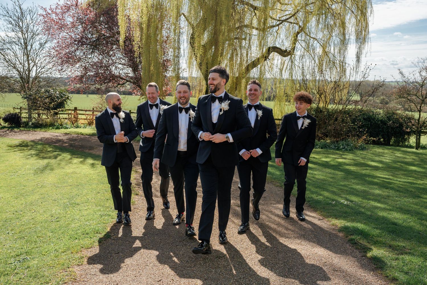 Groom and groomsmen in black tuxedos with bow ties walking together outdoors near a willow tree.