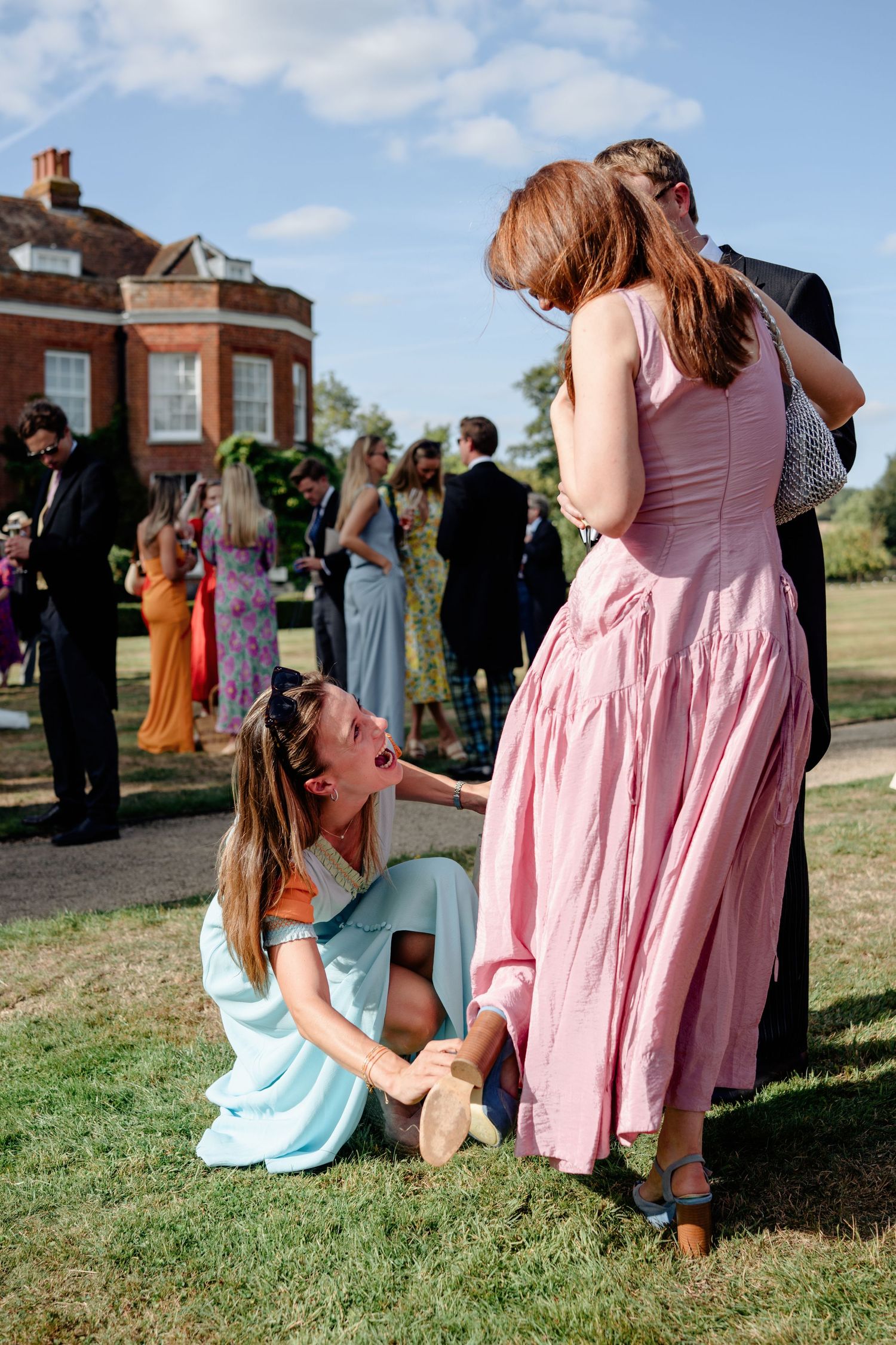 Wedding guest in blue dress sits on grass humorously grabbing friend's ankle at sunny garden party.