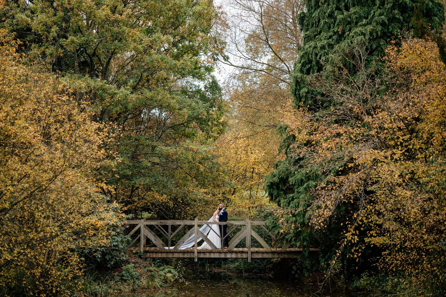 Bride and groom stand on wooden bridge surrounded by vibrant autumn foliage.