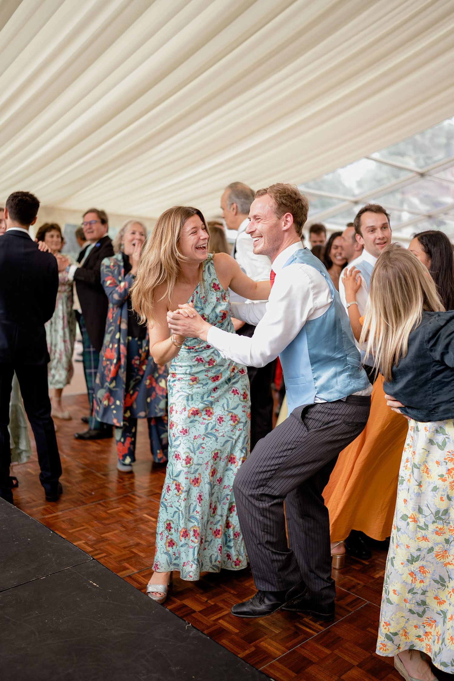 Wedding guests laughing and dancing energetically on a wooden dance floor inside a white marquee reception.