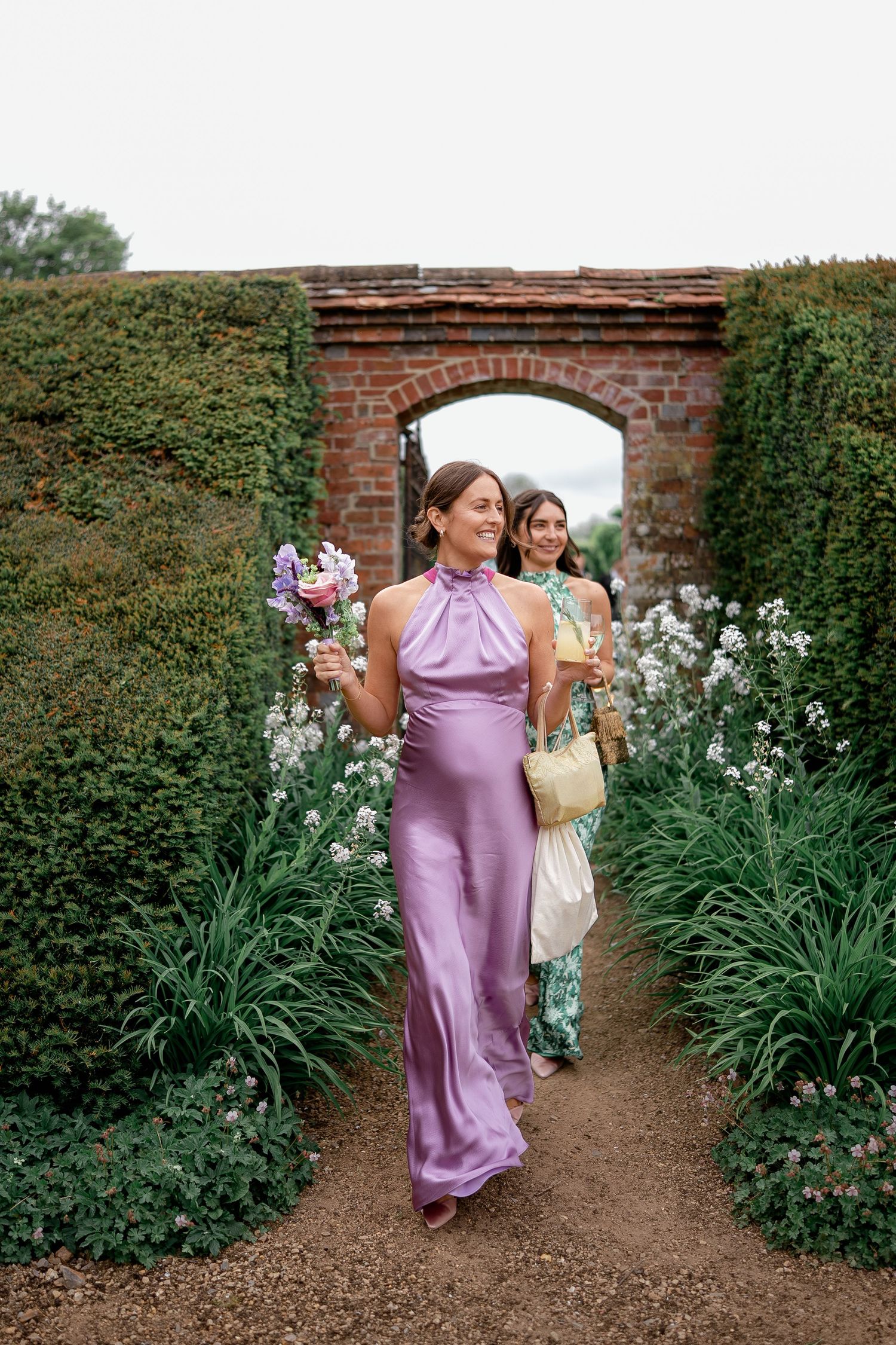Bridesmaid in lavender satin halter gown walking through a brick archway surrounded by lush garden greenery.