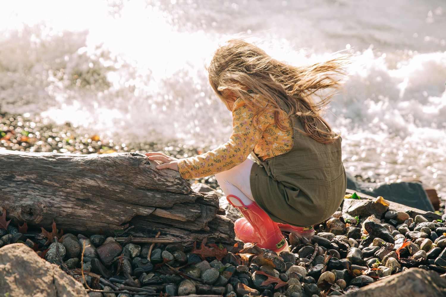 Young girl with windswept hair exploring rocky beach, reaching toward driftwood with ocean waves crashing behind her. Lincoln Park Seattle