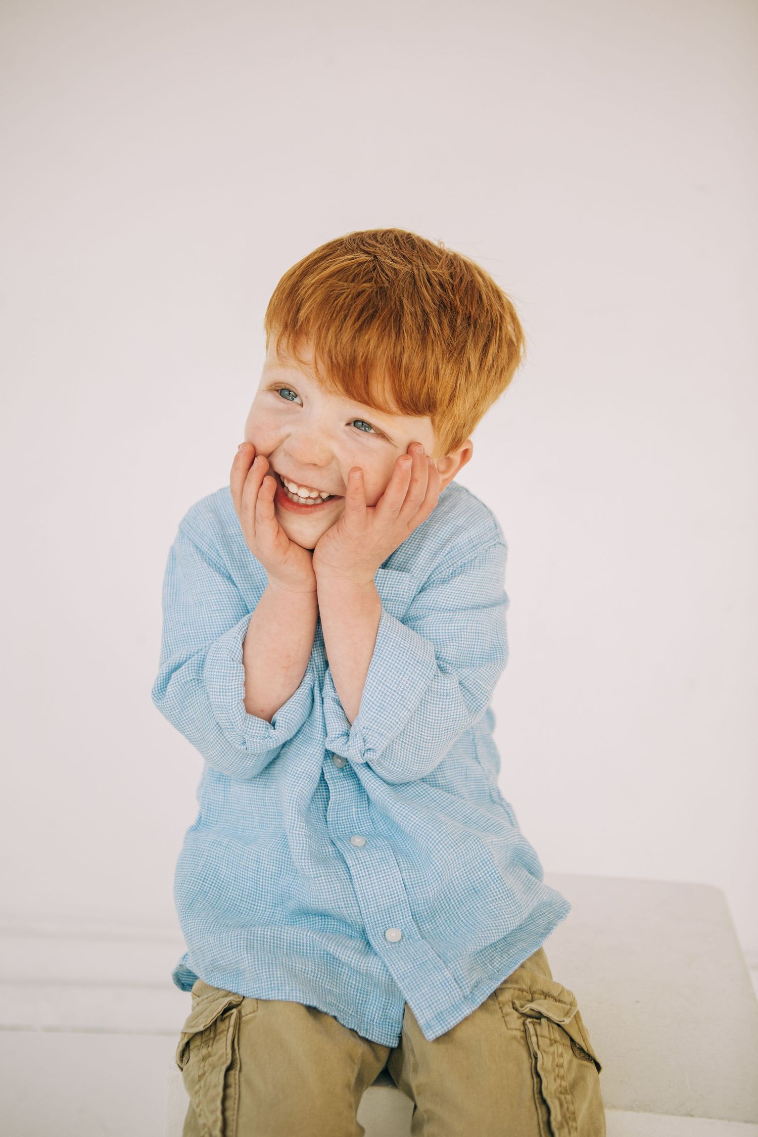 Adorable red-haired boy in blue shirt smiling with hands on cheeks against white background- Seattle, Washington; Child Photography Studio