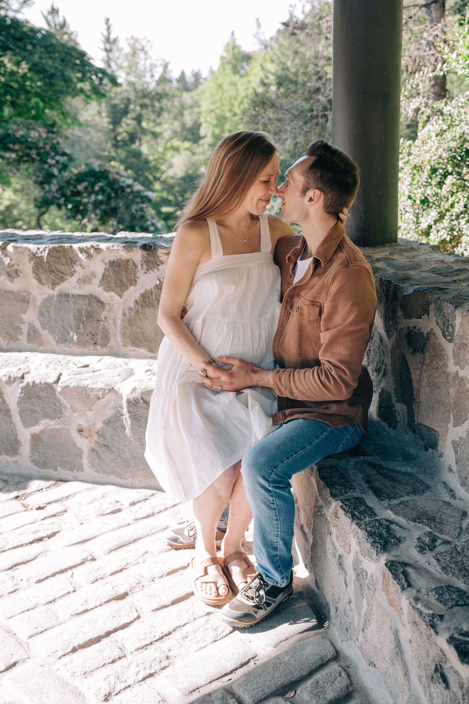Romantic couple sharing a tender moment on a stone terrace surrounded by lush green trees on a sunny day. in Seattle WA at Arboretum