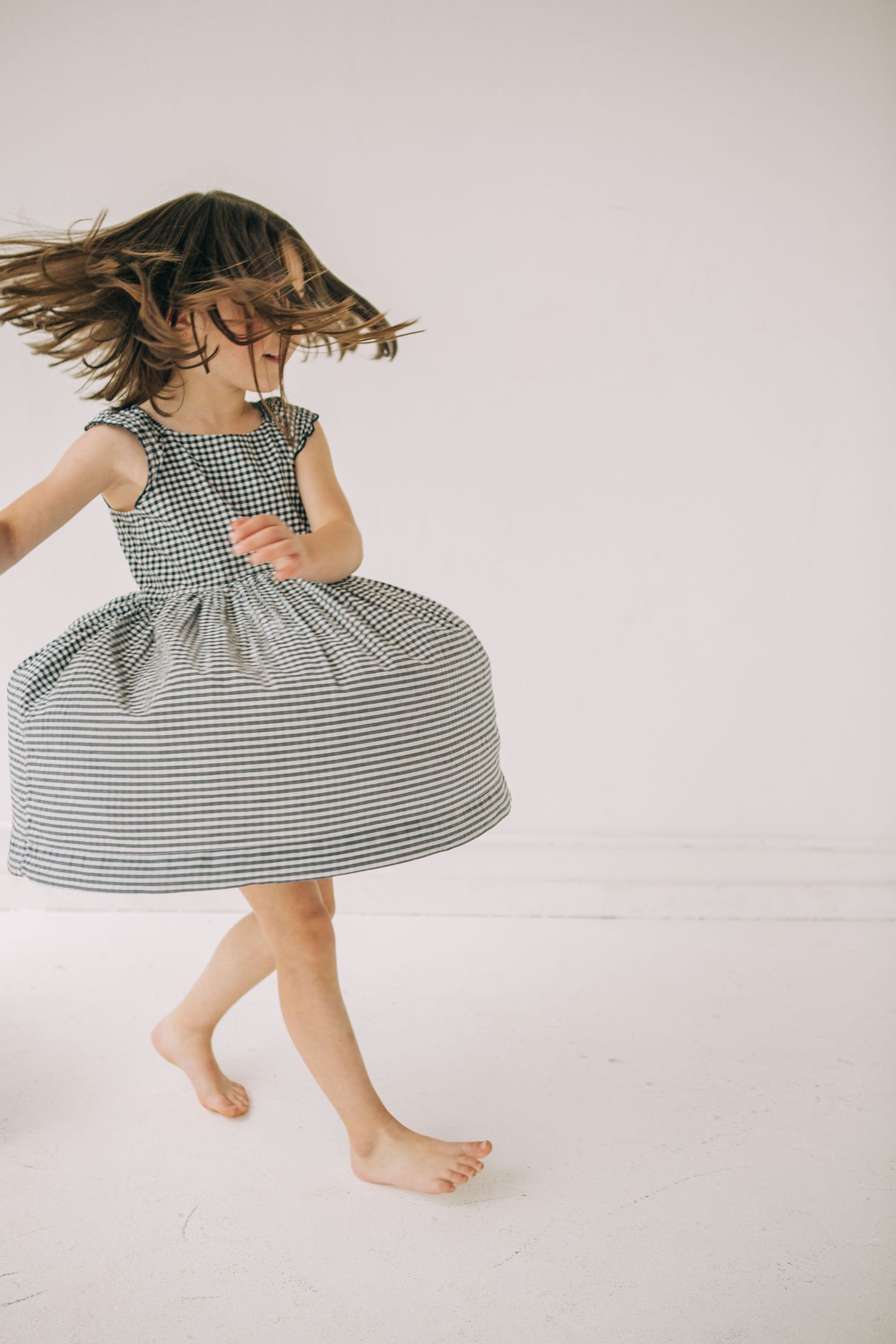A young girl in a gingham dress twirls barefoot, her hair flying as she dances joyfully in a bright white room. in Seattle Washington