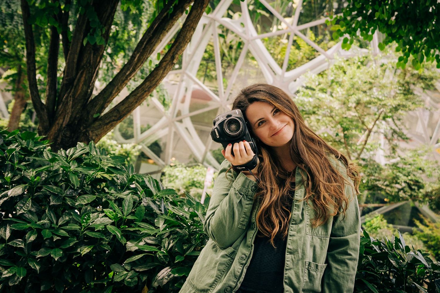 Young woman holding a DSLR camera outdoors, smiling among lush green plants with a white geodesic structure behind her in Seattle Washington