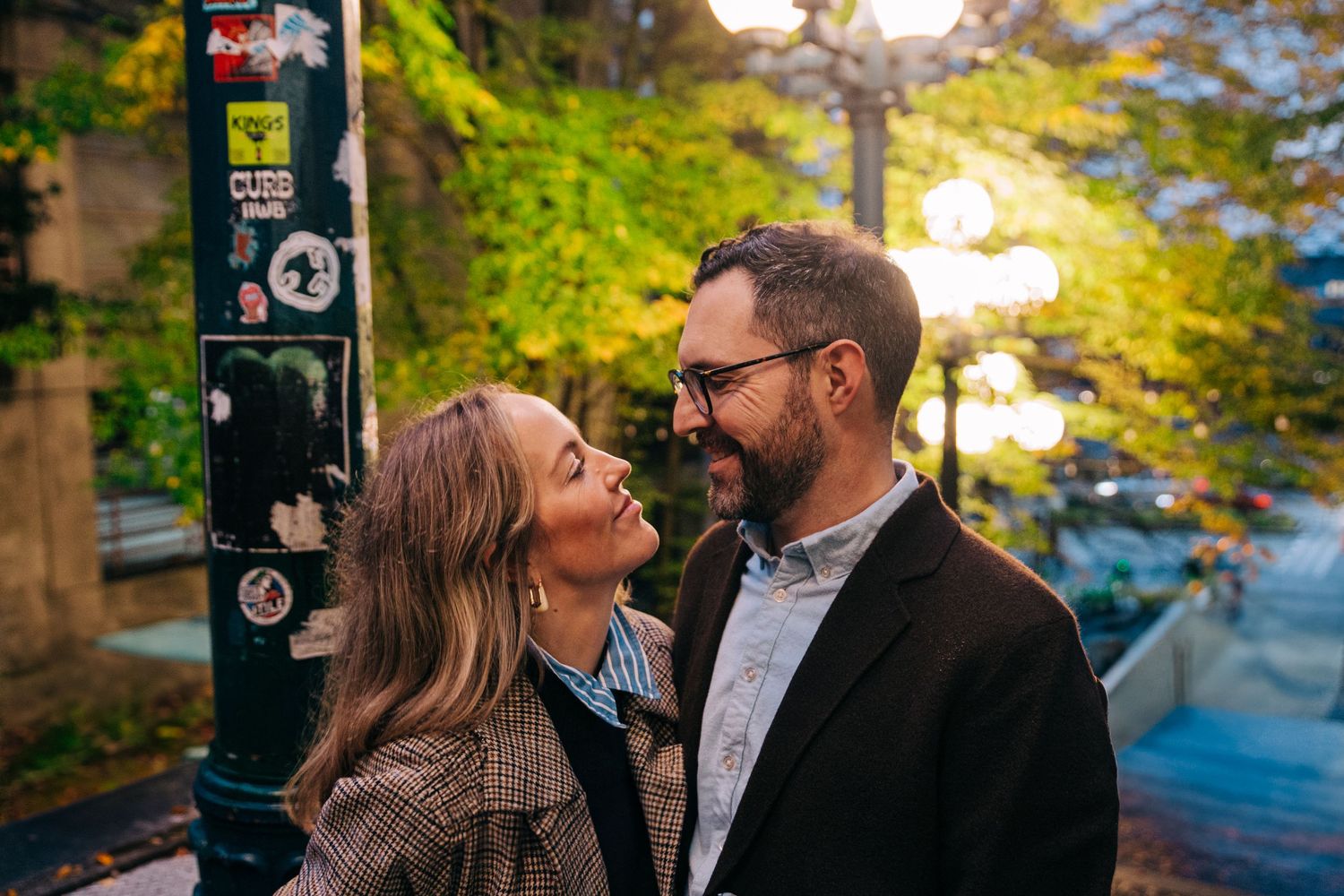 A couple shares a playful moment on a tree-lined urban street in autumn, the woman puckering her lips toward the smiling man. At Pikes Place