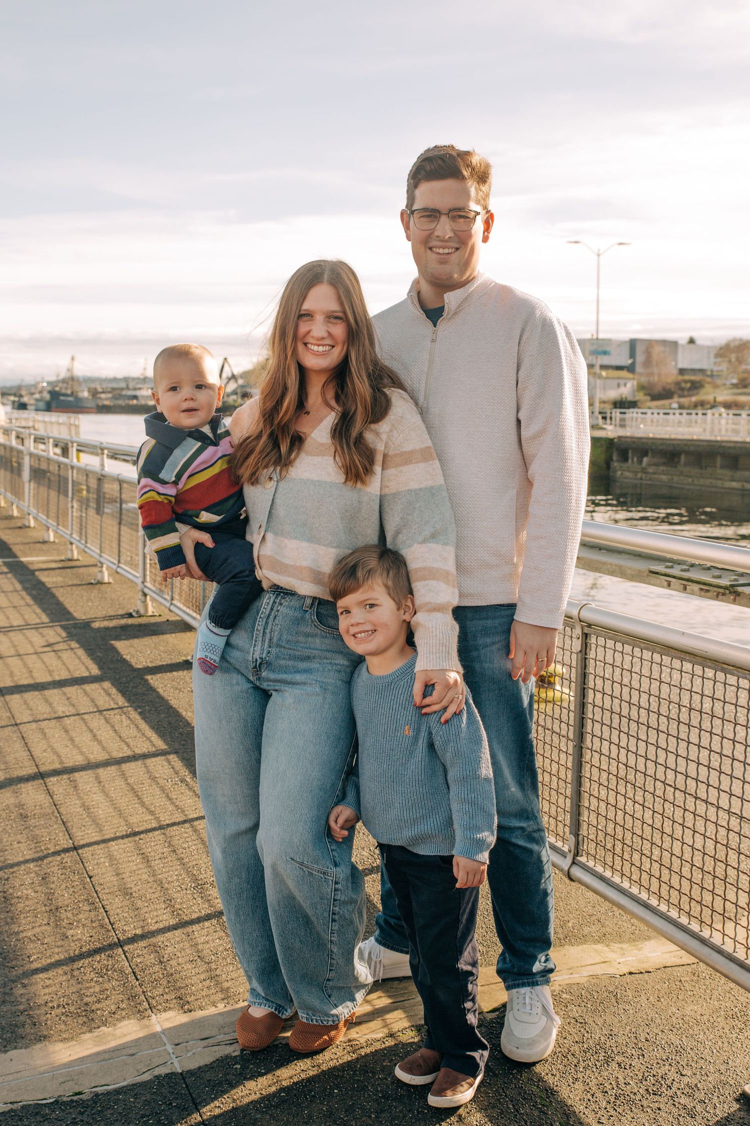 Happy family of four posing on a waterfront bridge on a sunny day, parents holding young children in casual attire. at Ballard Locks, in WA