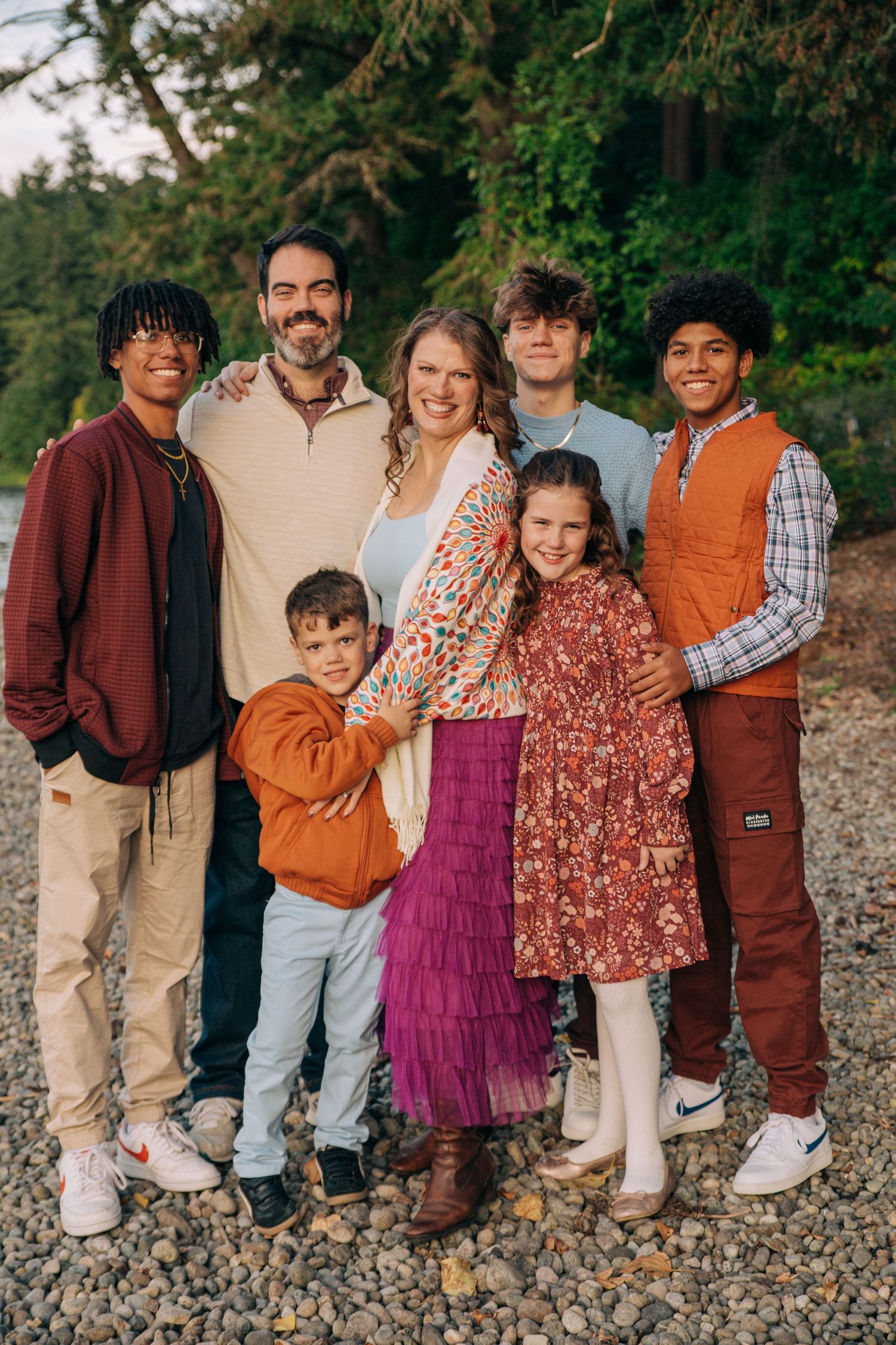 A joyful family of seven poses together on a rocky beach surrounded by lush green trees. In seattle Washington Seward Park