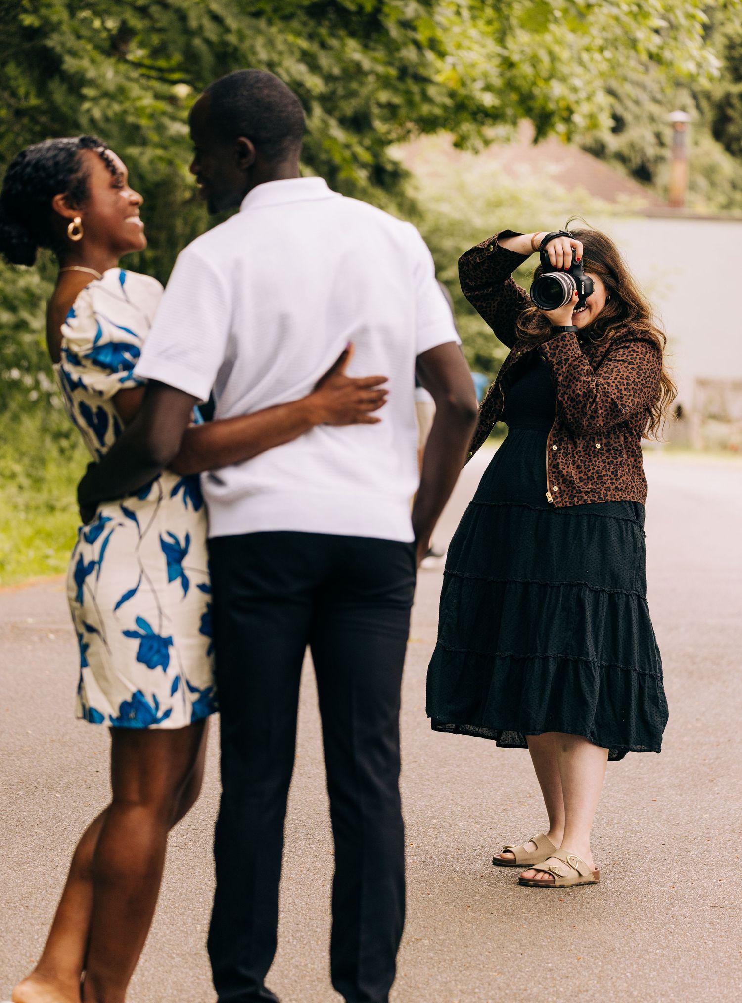 A photographer captures engaged couple embracing outdoors on a path surrounded by lush green trees @ Seward Park, Seattle WA