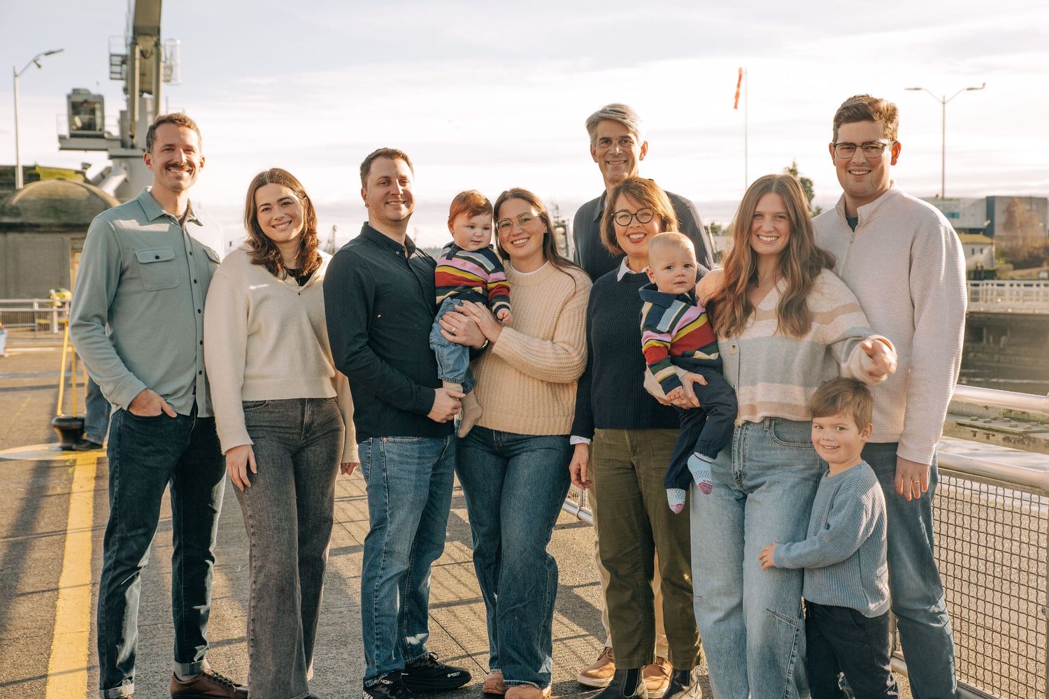 A large family group smiling together outdoors on a bridge at sunset, dressed in casual coordinated outfits. At Ballard Locks, in Seattle WA