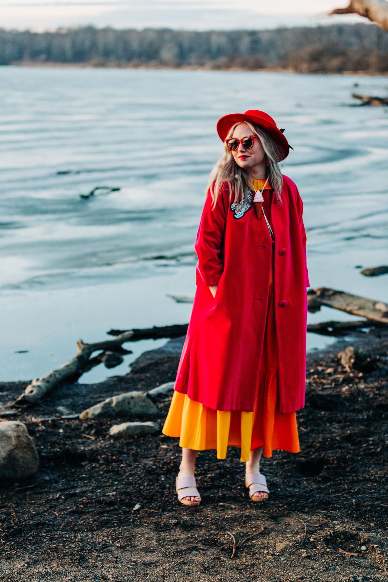 Woman in red coat, hat, sunglasses, and yellow dress stands on rocky shore near water. In Seattle Washington