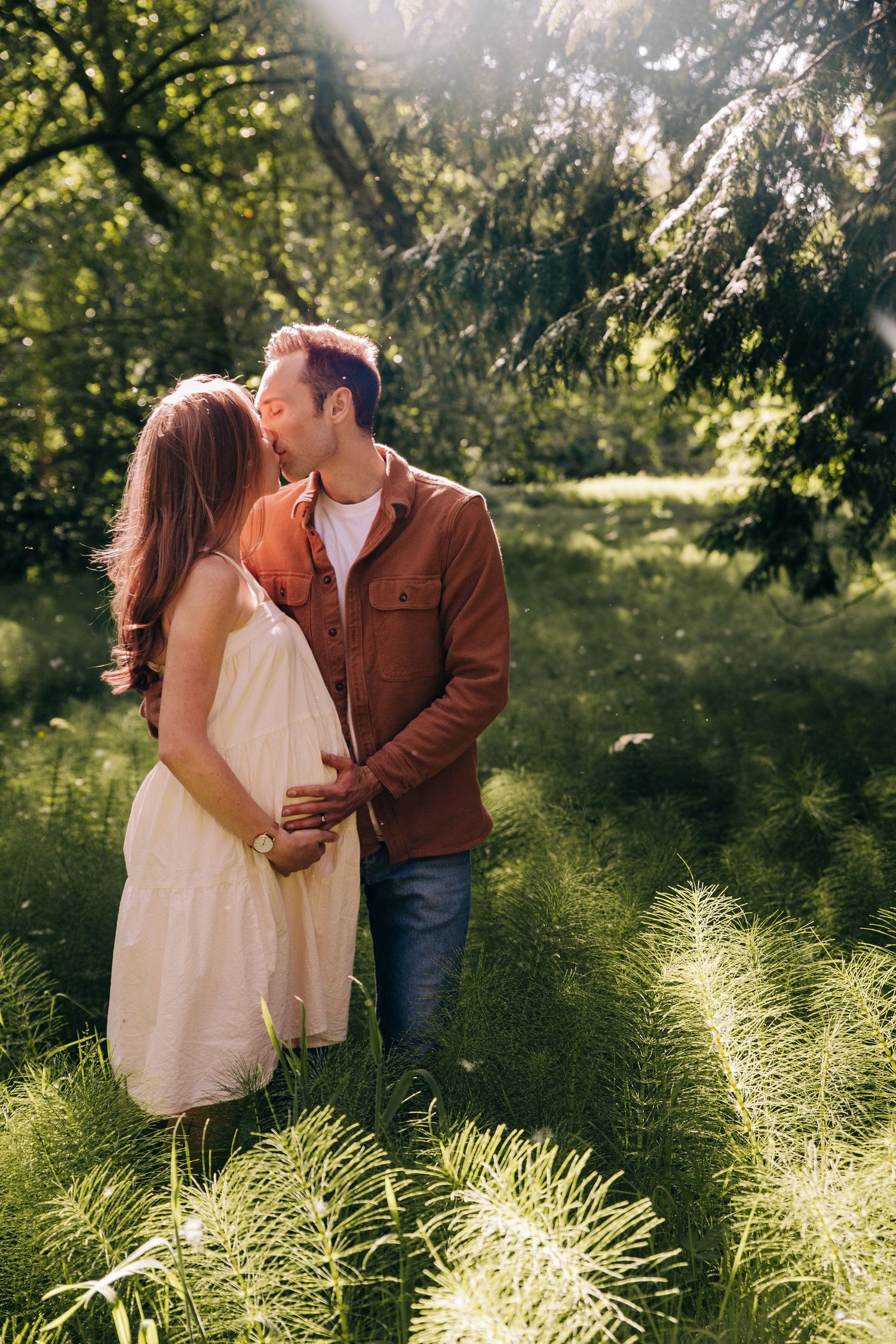 A couple shares a tender kiss in a sunlit green garden, the woman in a white dress cradling her pregnant belly. In seattle washington