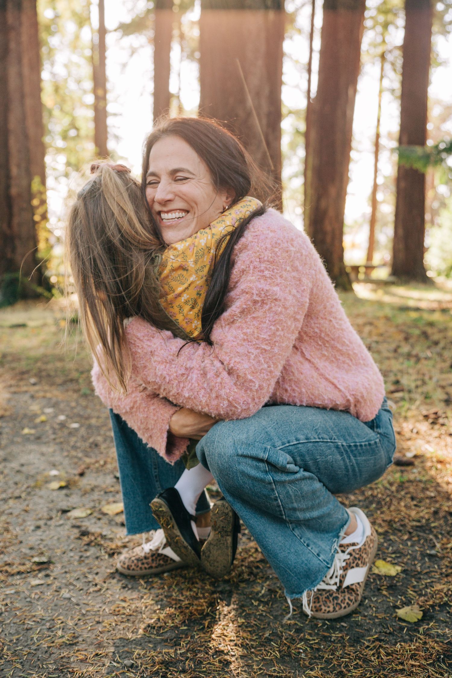 Woman in pink sweater crouching to hug a young girl in a sunlit forest setting. At Lincoln Park in Seattle, Washington. Family Photo Session