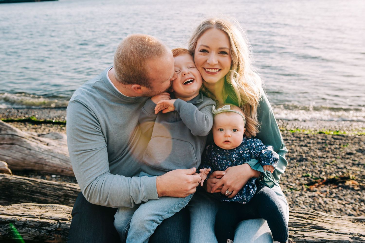 Happy family of four sitting by the water, parents smiling with toddler and baby on a rocky beach at Lincoln Park, Seattle WA