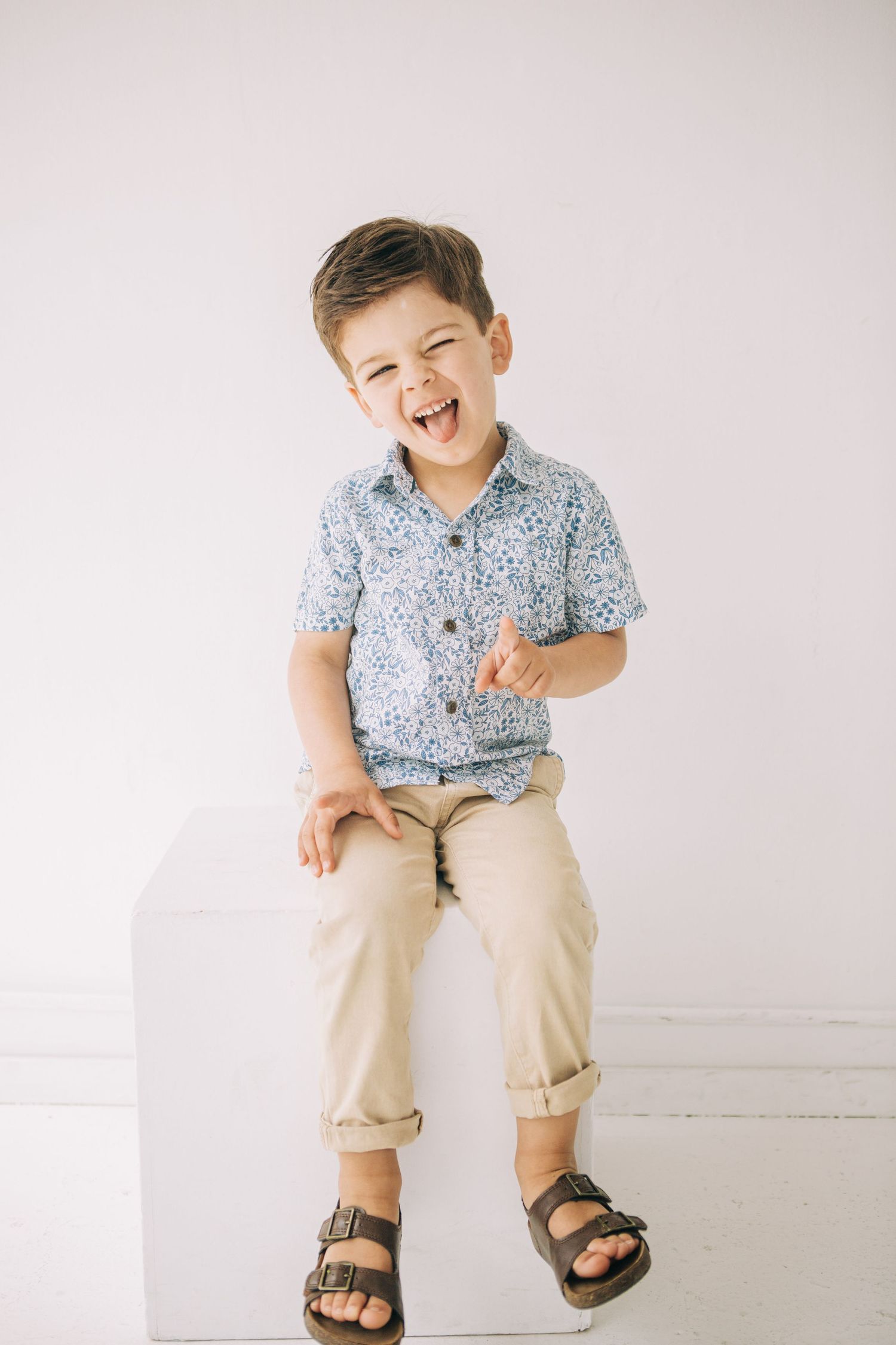A laughing young boy sits on a white cube wearing a floral shirt, khaki pants, and brown sandals. in Seattle Washington at Children's Studio