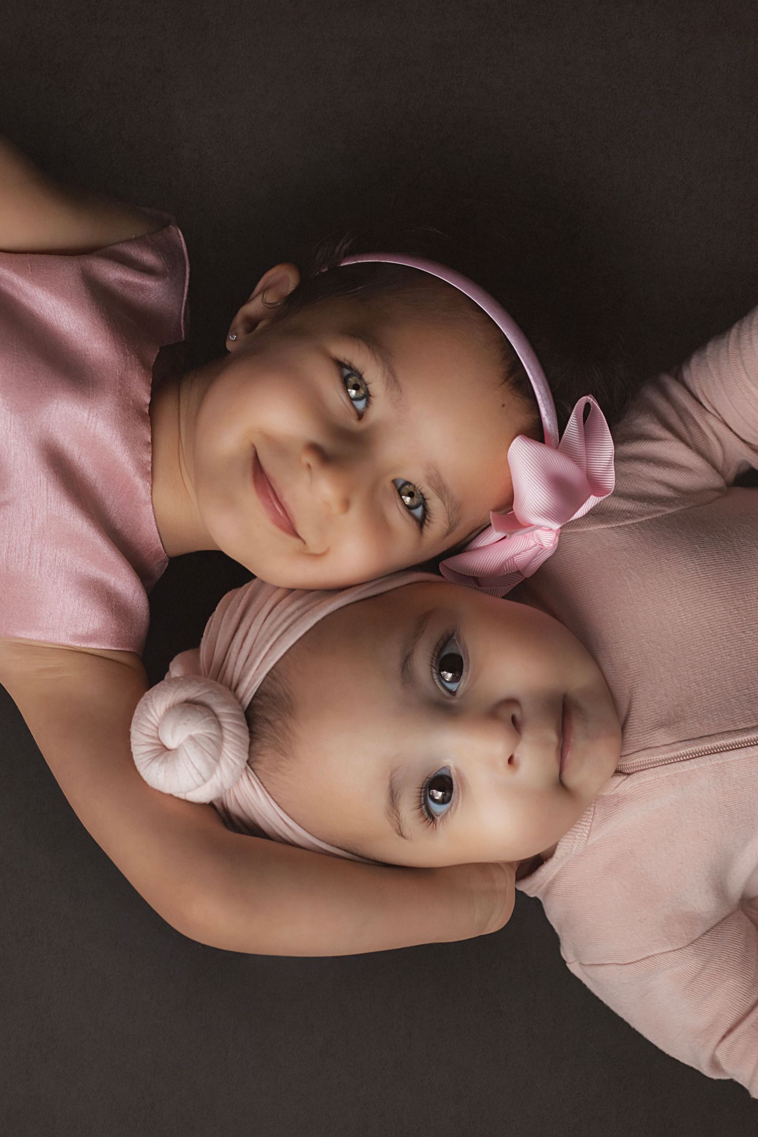 Two young sisters in coordinated, pink outfits lying close together in a fine art sibling portrait with soft, dreamy styling.
