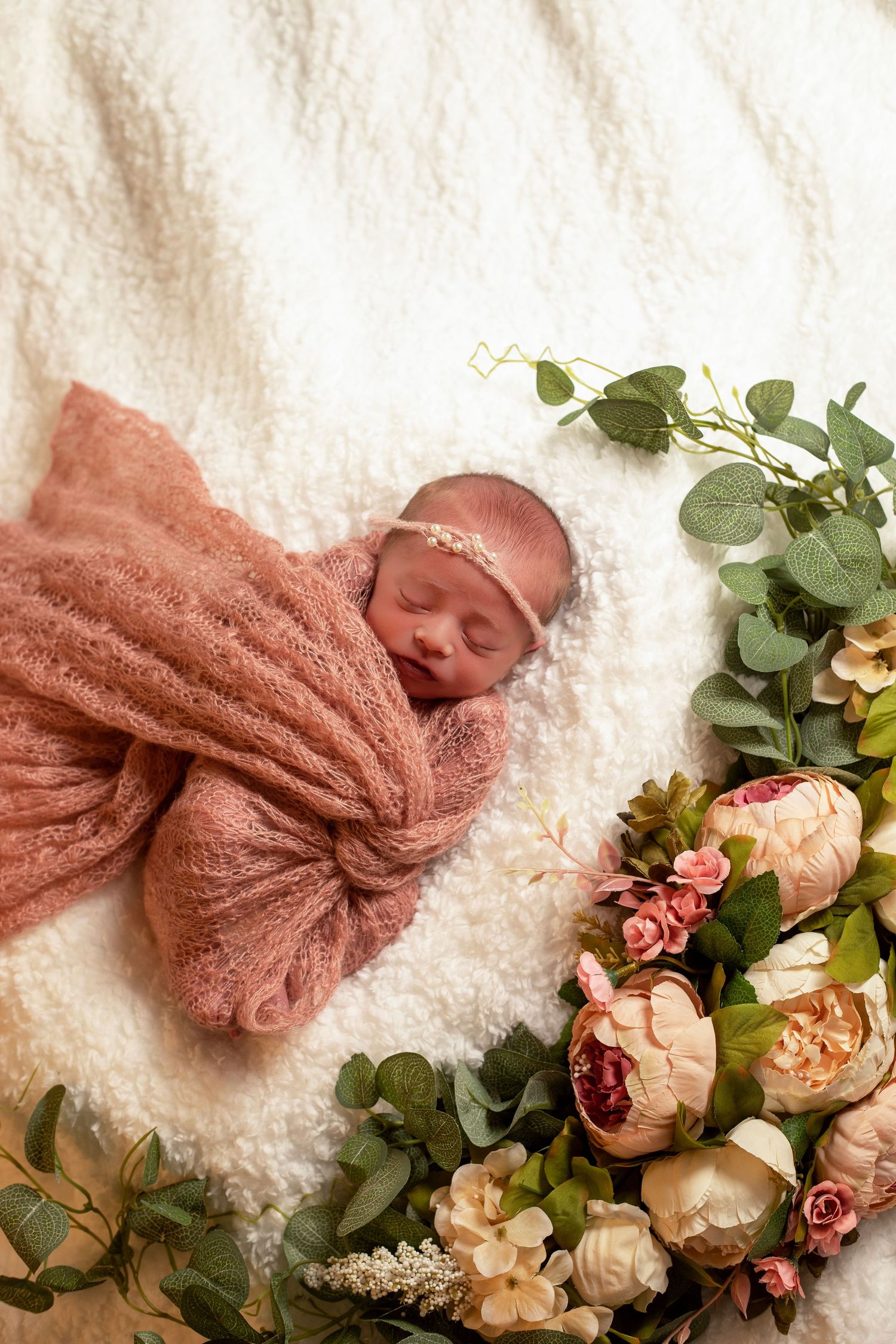 Newborn baby wrapped in blush fabric, beside flowers in a fine art, newborn portrait with soft natural tones.