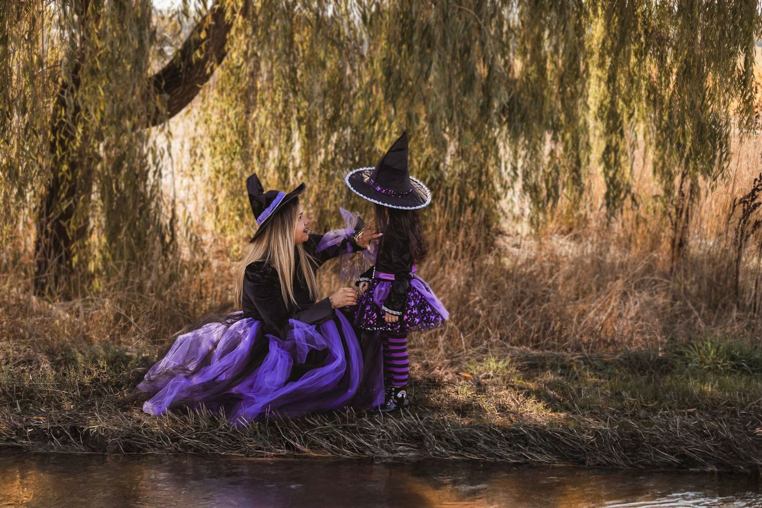 Whimsical which inspired portrait featuring a mother and daughter in fantasy costumes beside a reflective pond under a willow tree