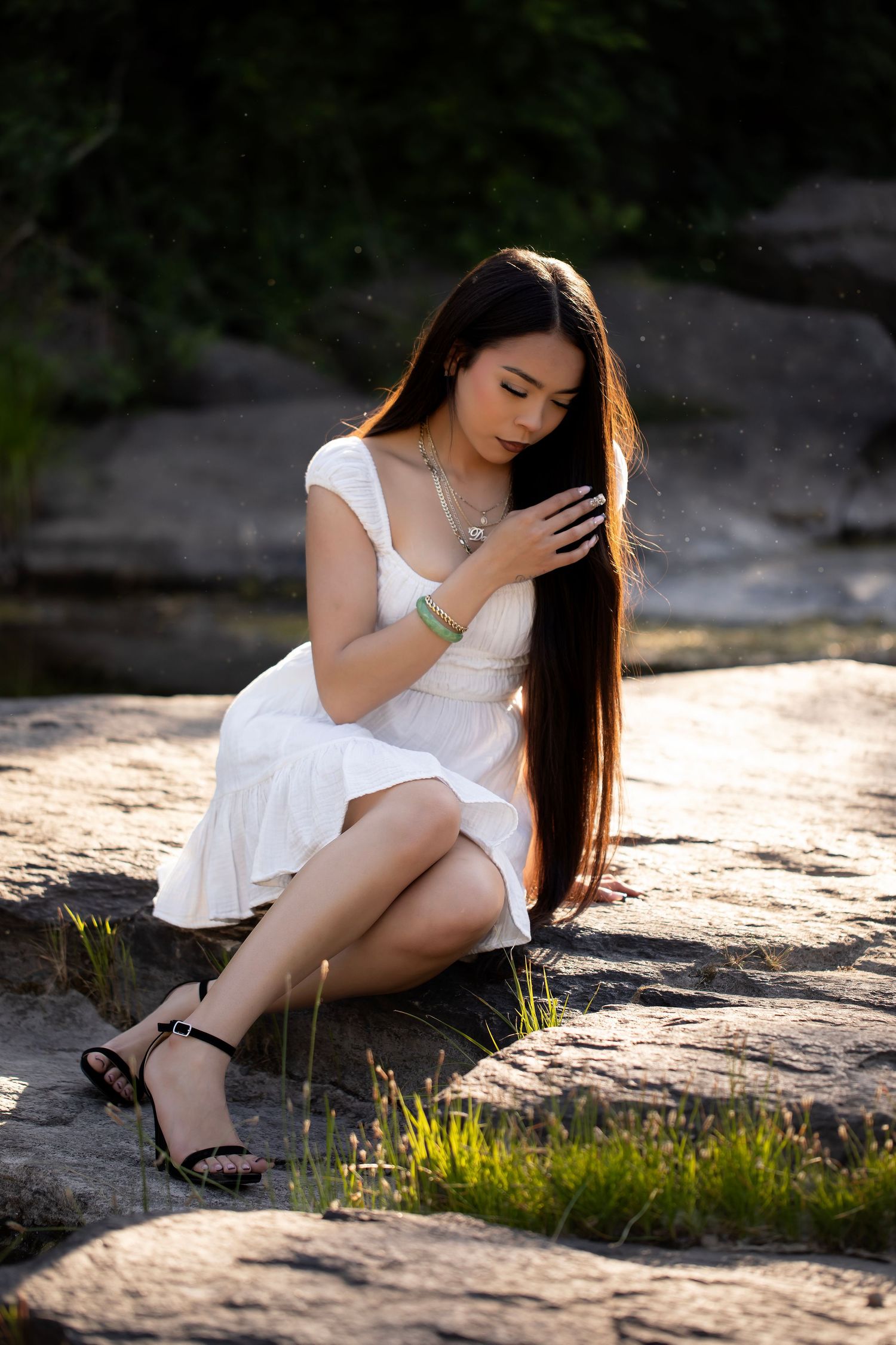 Fine art portrait of a high school senior seated on riverside rocks wearing a flowing white dress in natural light