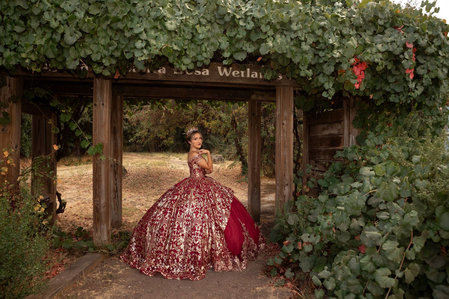 Outdoor quinceañera portrait featuring a young woman, and a richly detailed ballgown framed by a garden arbor