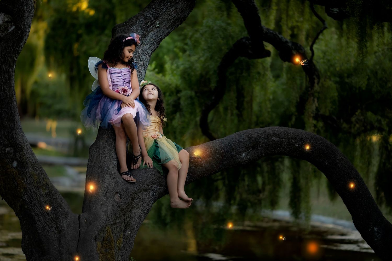 Fantasy childhood portrait featuring sisters seated in a tree surrounded by glowing firefly-inspired light effects