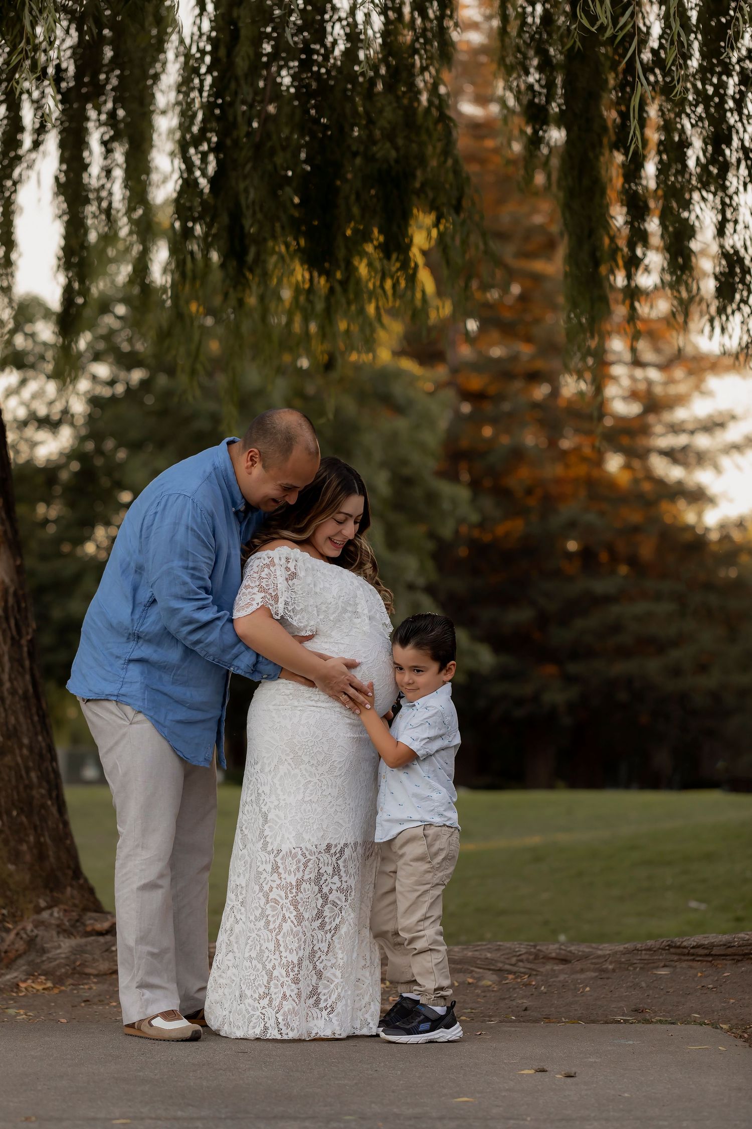 Family maternity portrait featuring parents and young child embracing beneath golden evening light