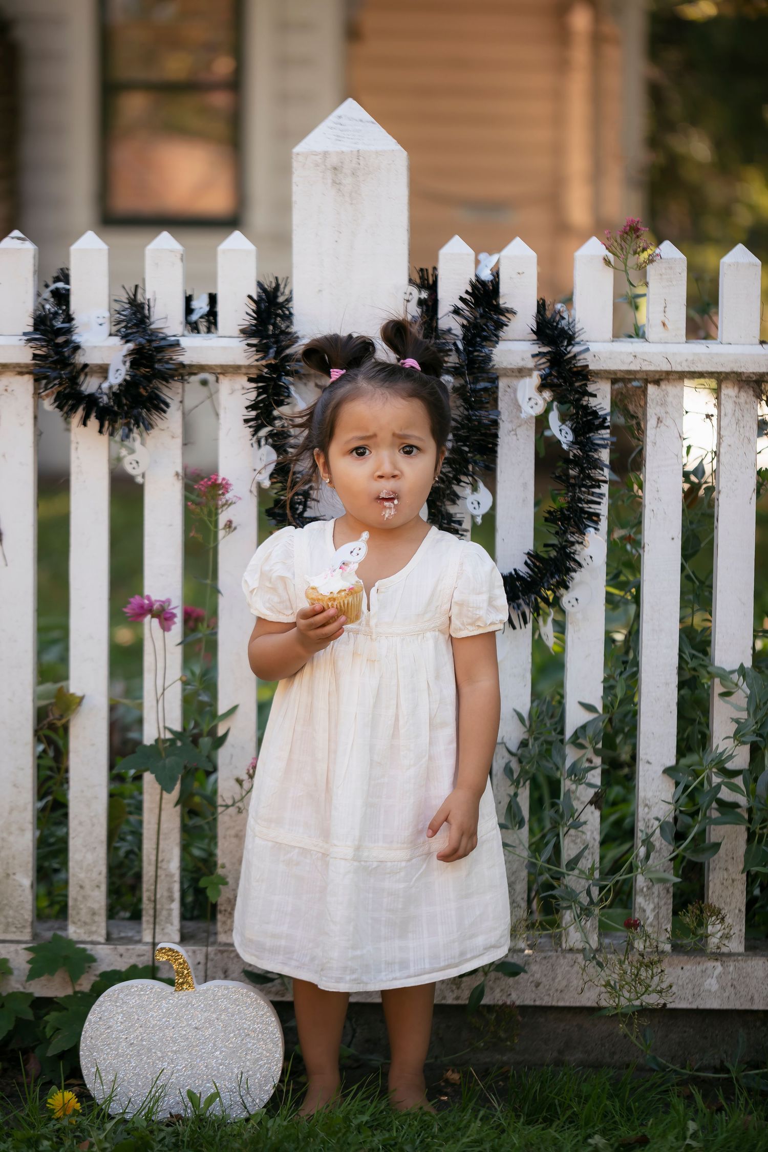 Fine art child portrait featuring a toddler in a white dress standing beside a picket fence and seasonal pumpkin decor