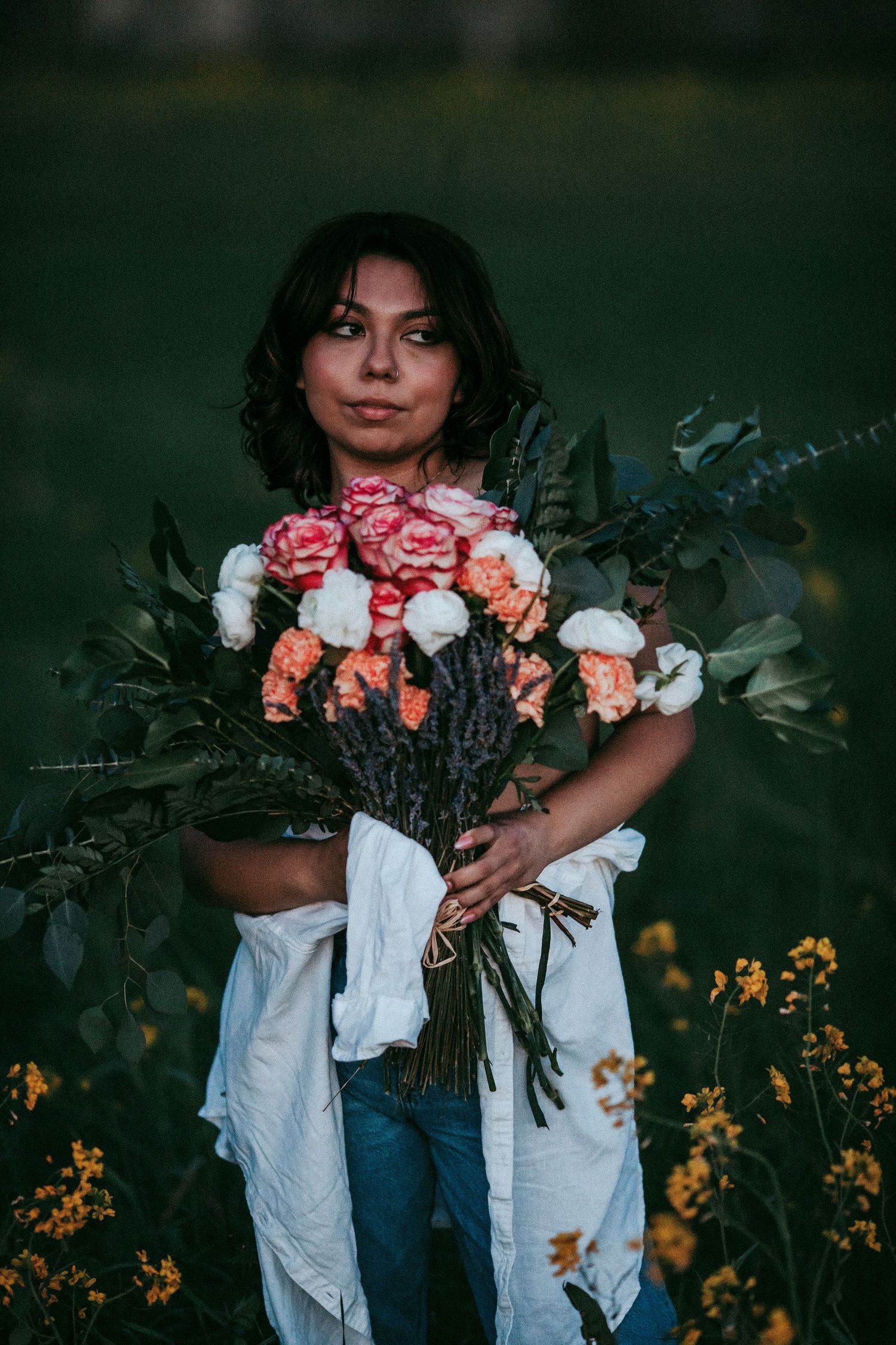 Fine art portrait of a young woman holding a bouquet of flowers, surrounded by wildflowers and a Moody garden setting