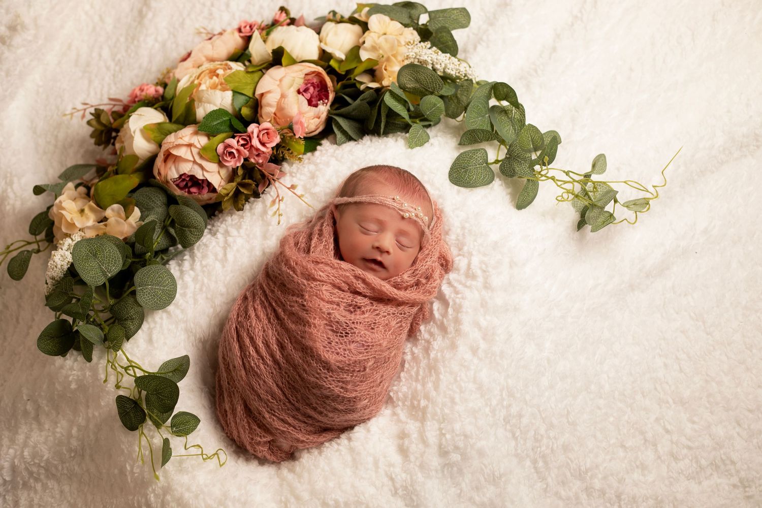 Newborn baby wrapped in blush fabric, beside flowers in a fine art, newborn portrait with soft natural tones.
