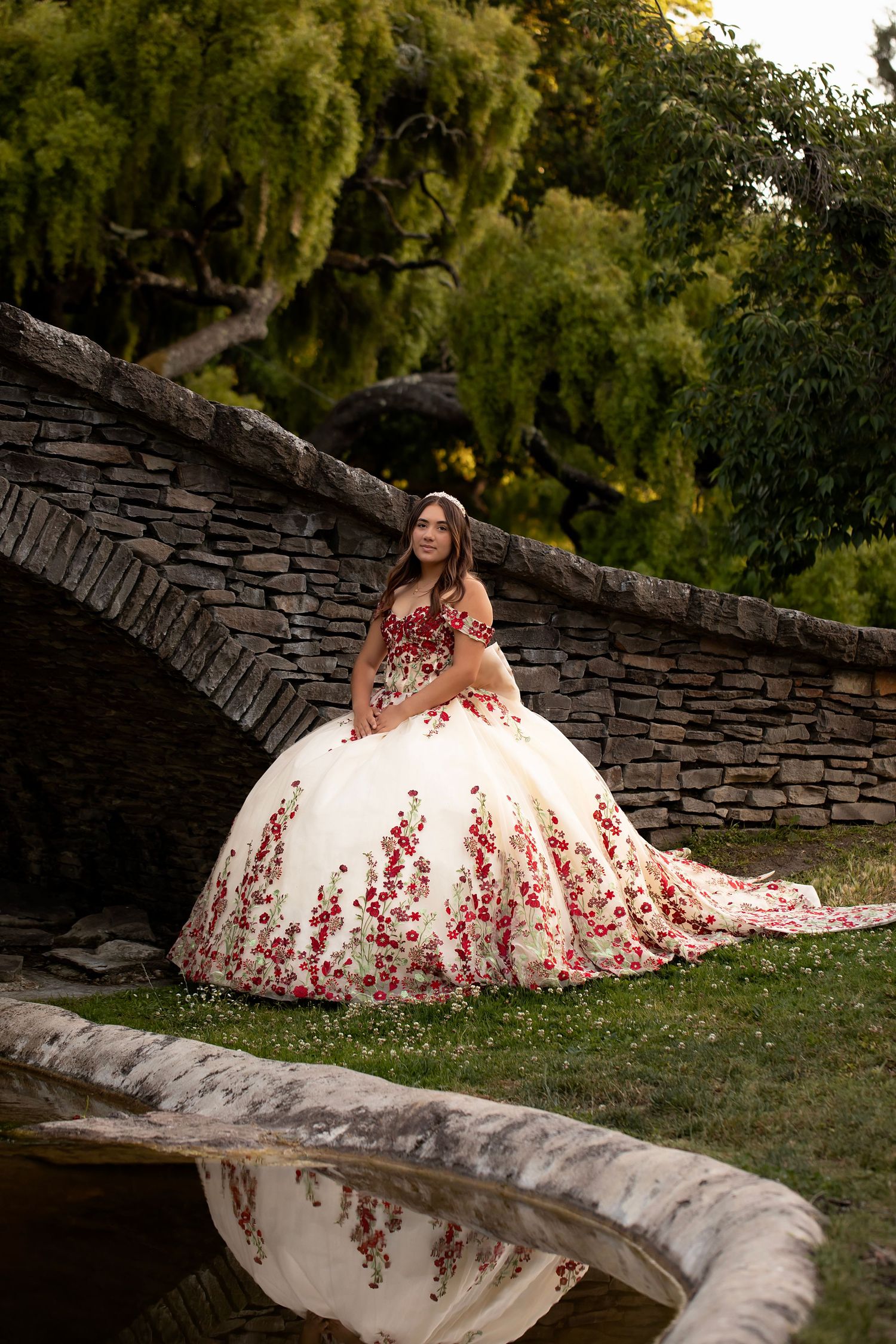 Outdoor quinceañera portrait featuring a young woman in a floral ball gown photographed beside a stone bridge and reflective water