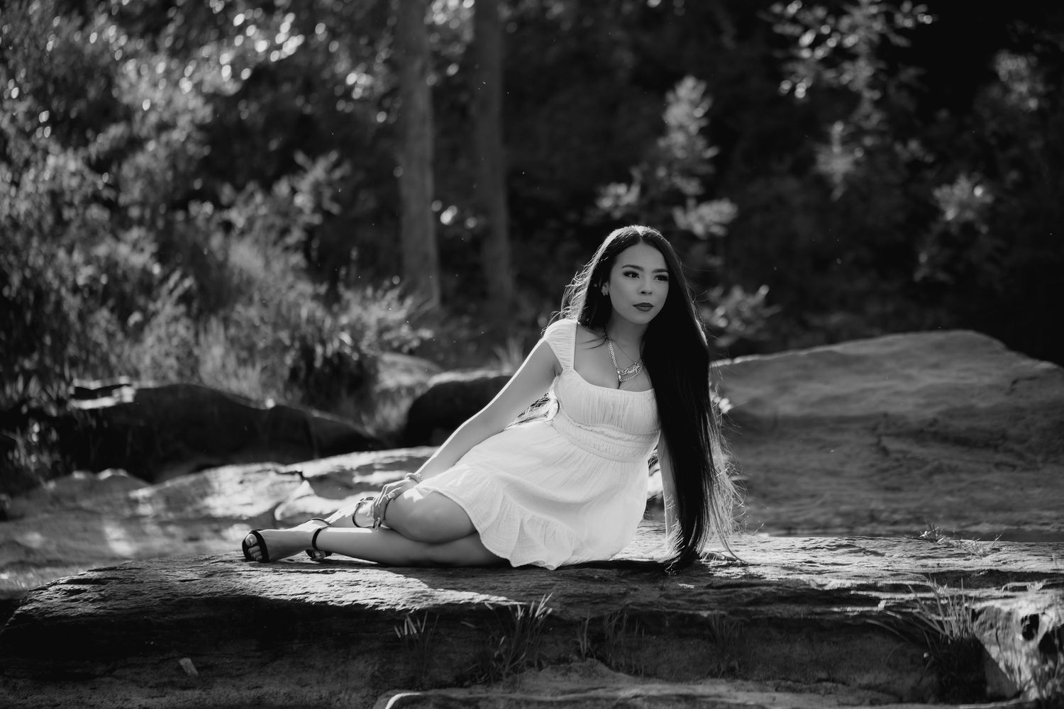 Black and white outdoor portrait featuring a high school senior in a white dress posed beside a calm stream