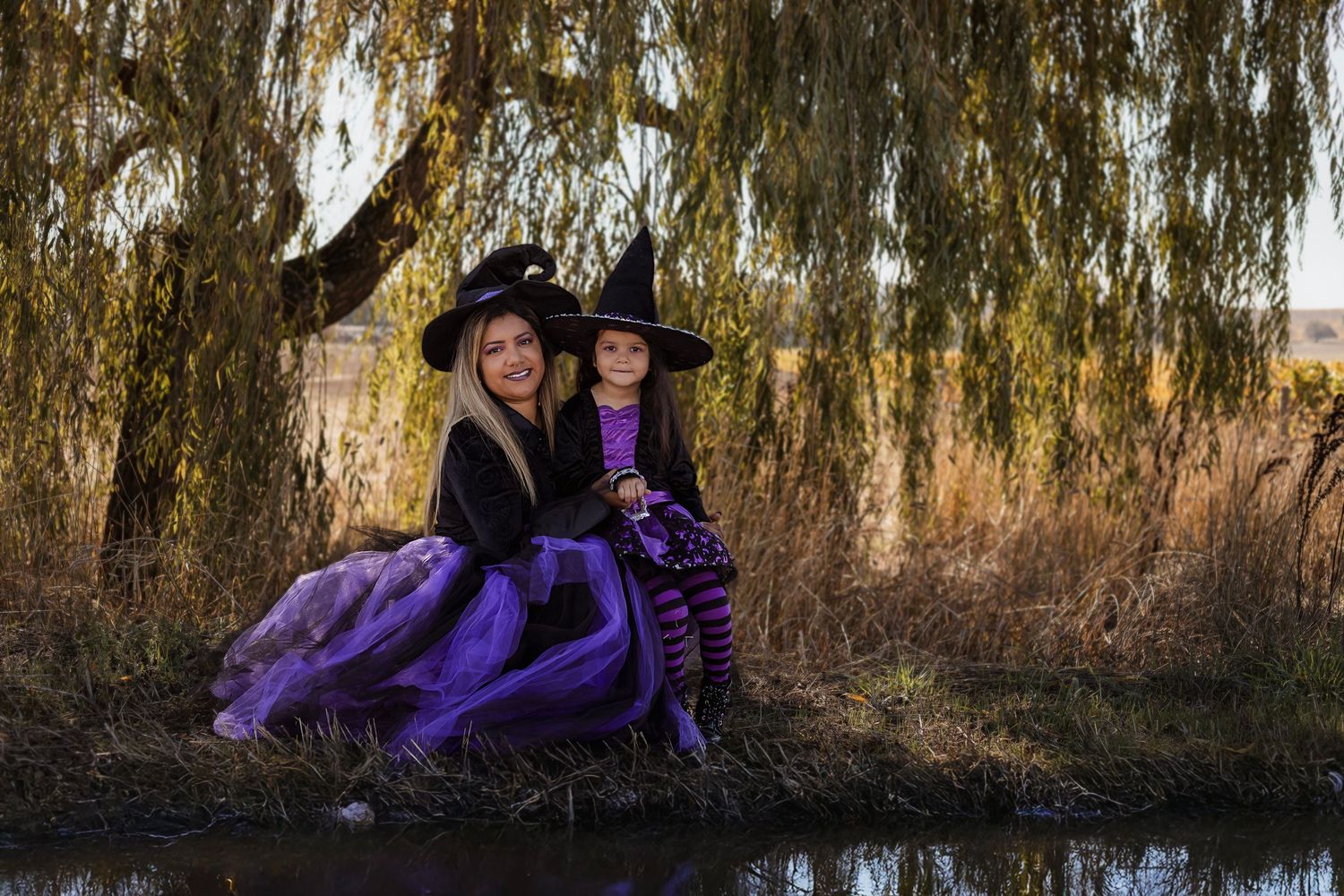 Fantasy portrait of a mother and daughter dressed as witches in flowing purple gowns beneath a willow tree beside a river