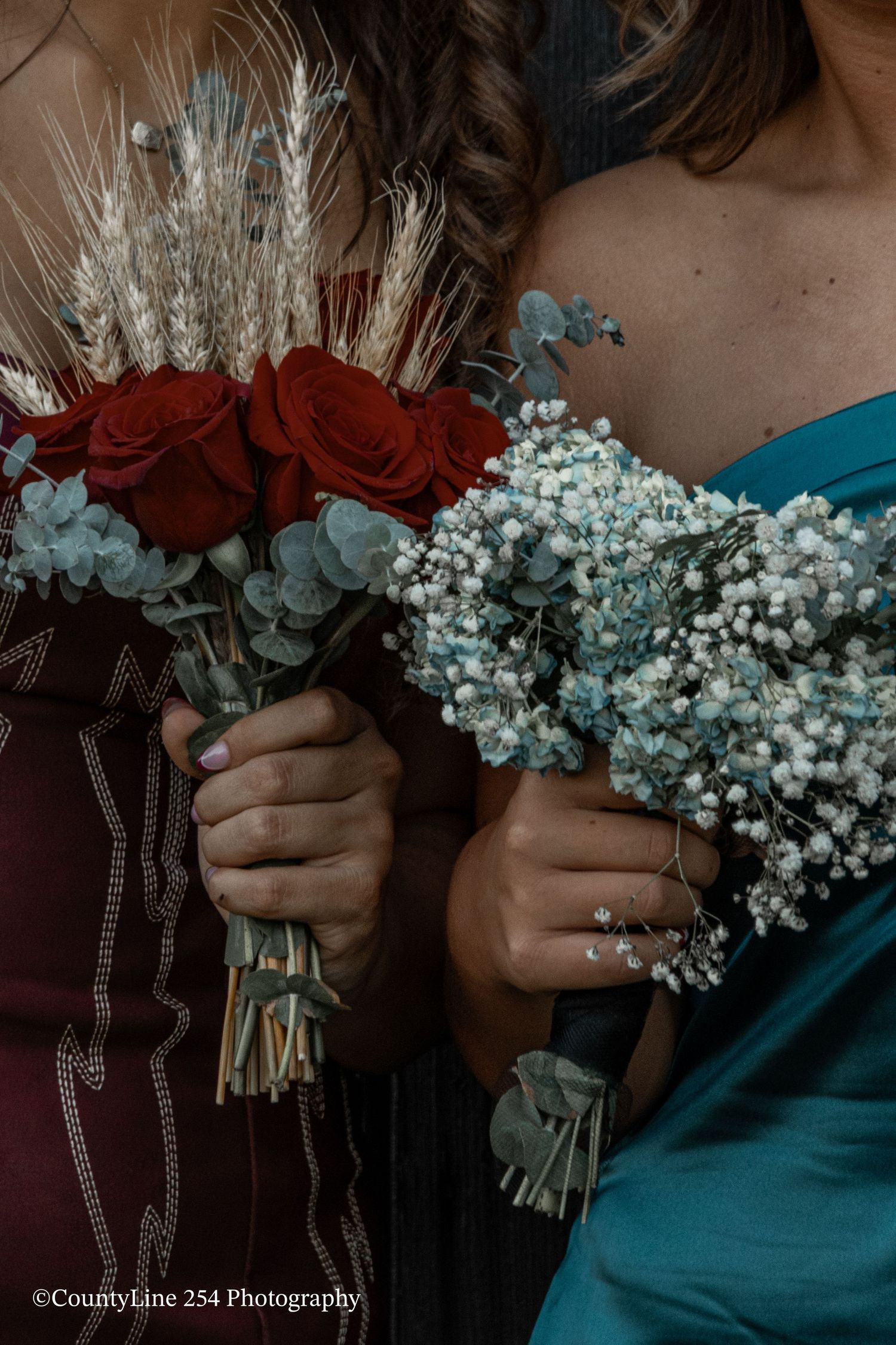Two women holding bridal bouquets of red roses and blue baby's breath in formal dresses.