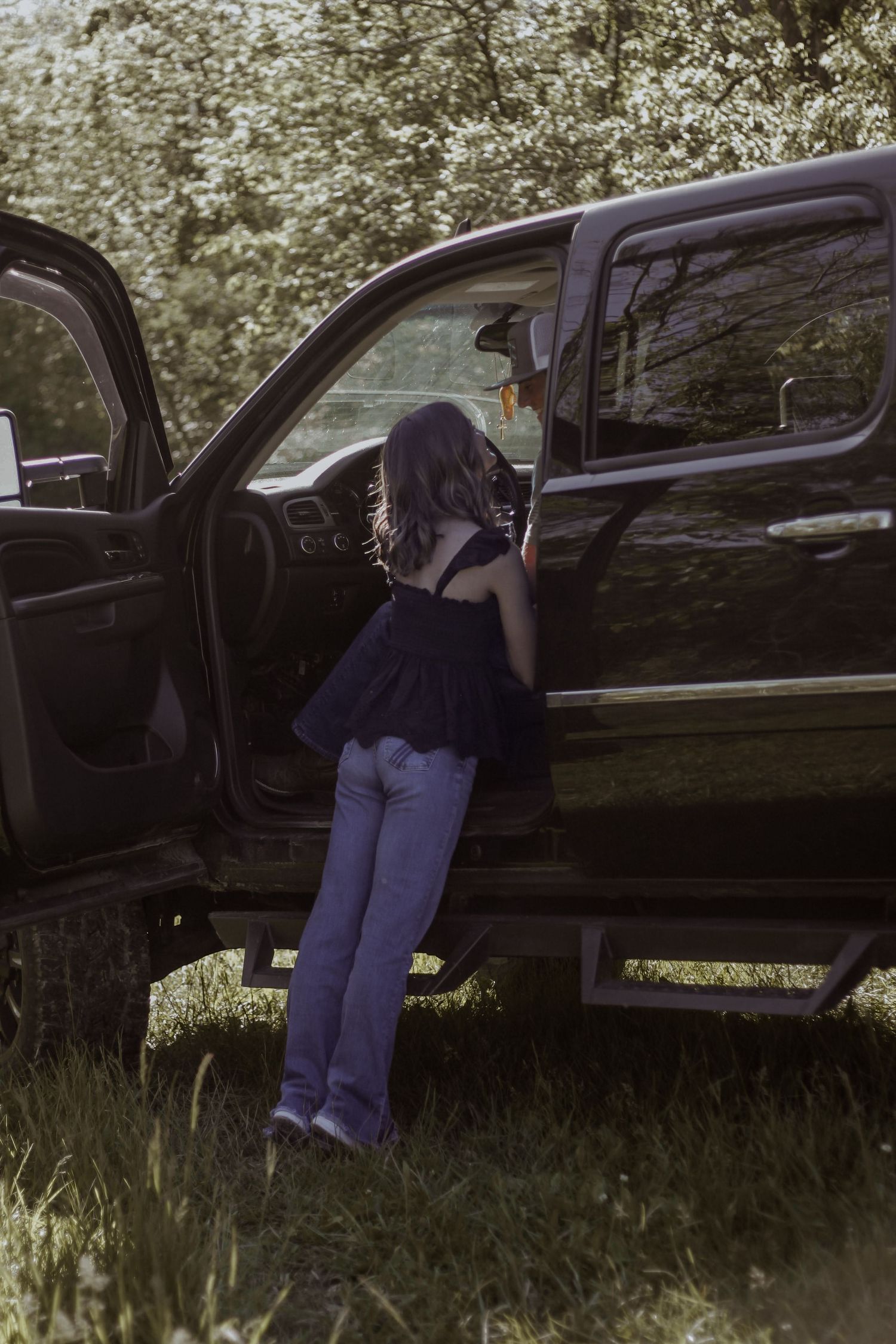 A young woman in a black top and jeans climbs into a large black pickup truck outdoors.