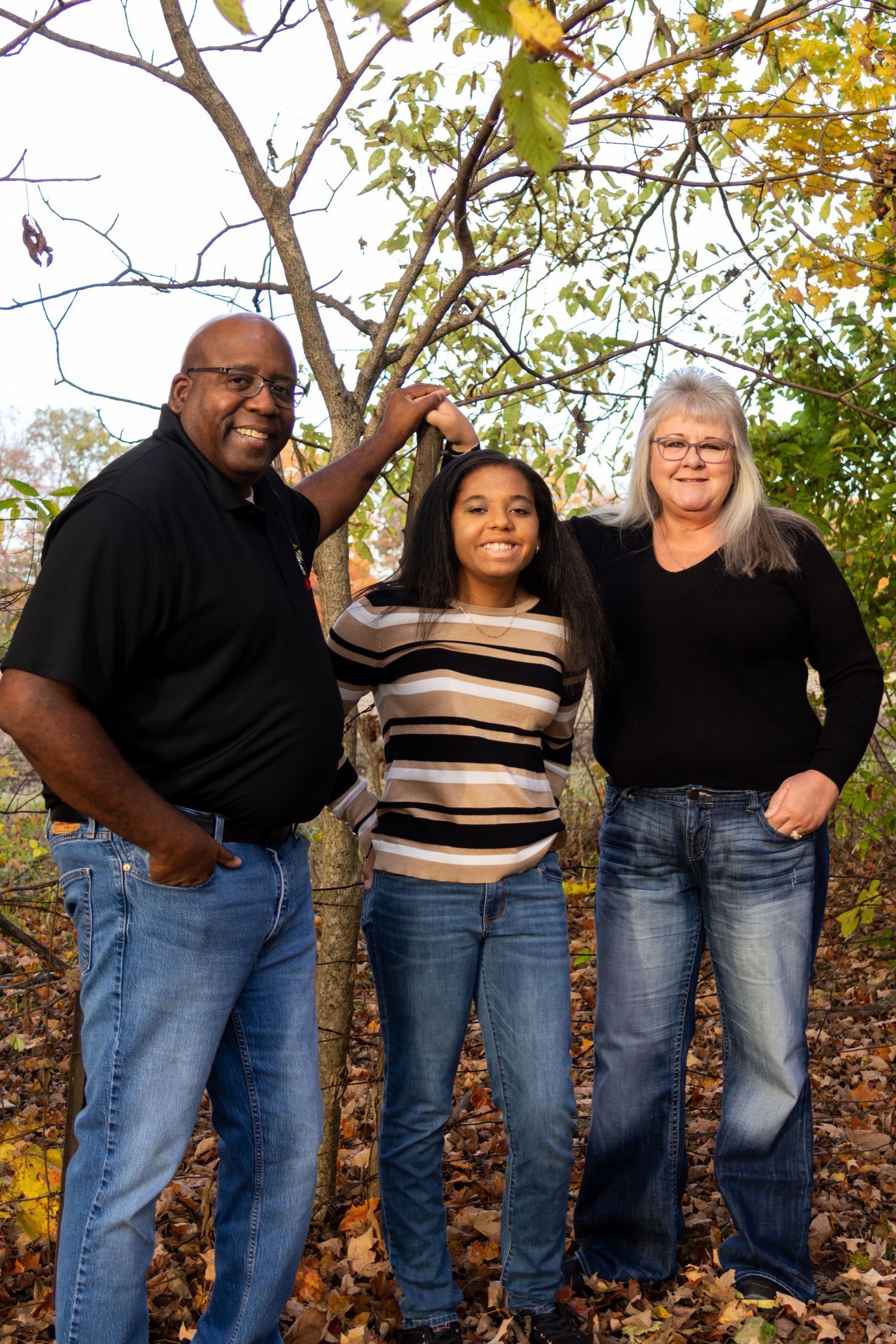 Smiling family of three standing under a tree in an autumn park, surrounded by fallen leaves and colorful foliage.
