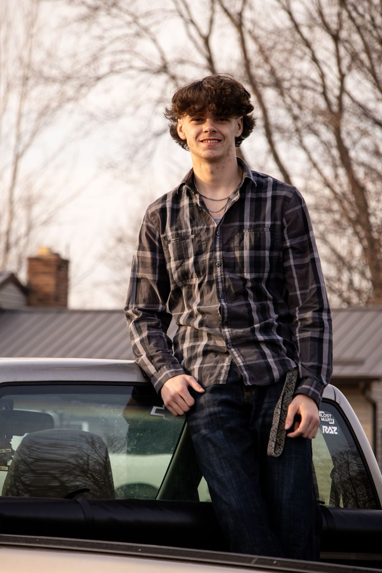 Young man in plaid shirt leaning against a silver car outdoors during senior portrait session.