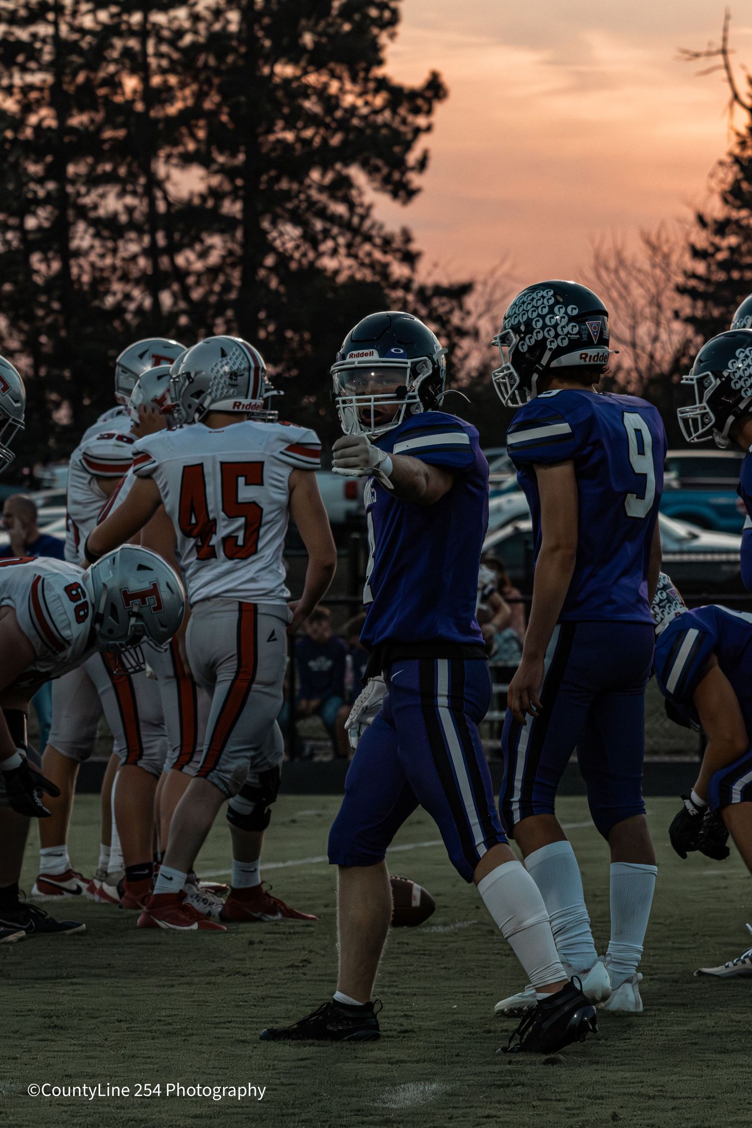 High school football players line up at scrimmage during a sunset game with player #45 visible.