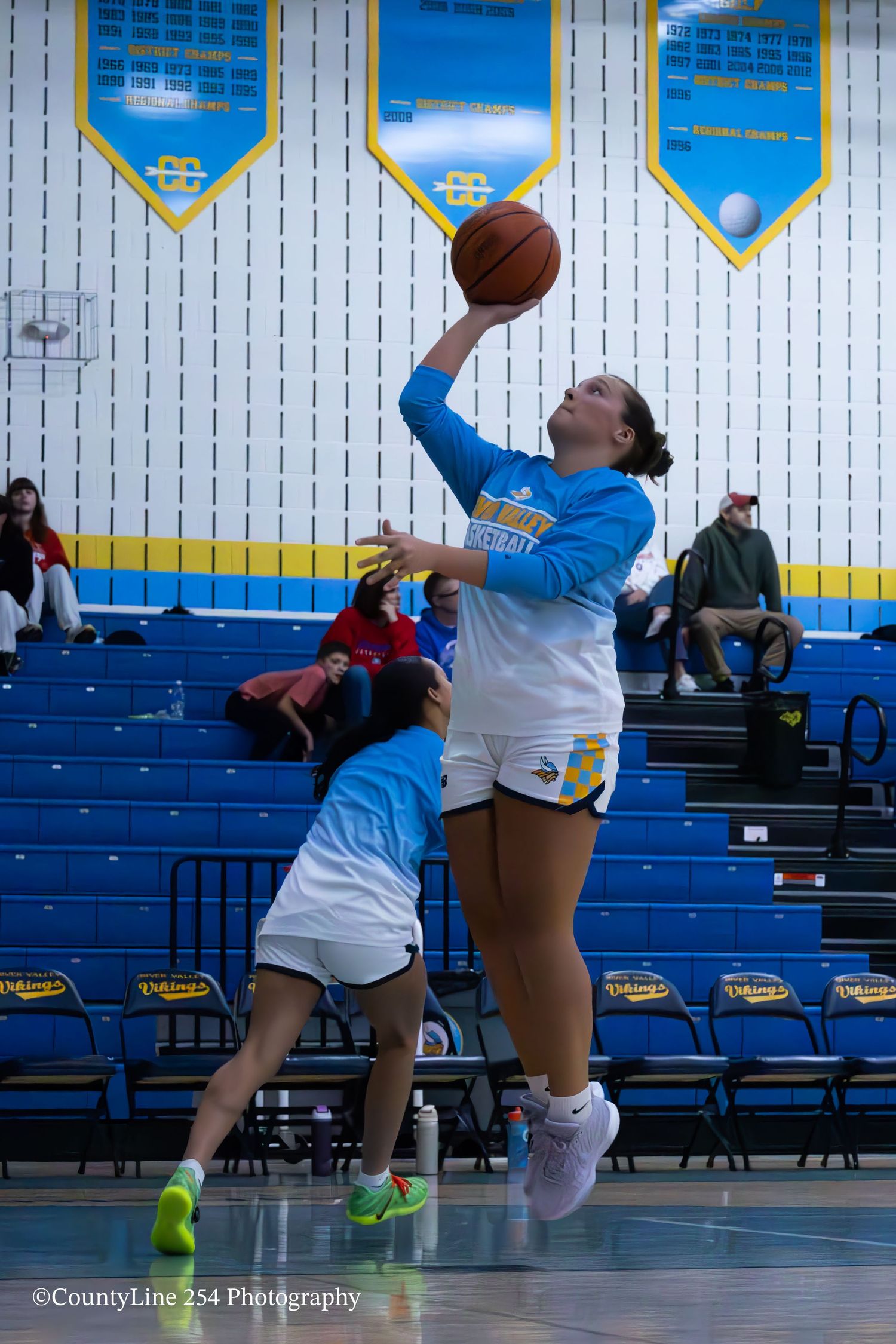 Two female basketball players practice a layup drill during warm-ups in a blue and yellow gym.
