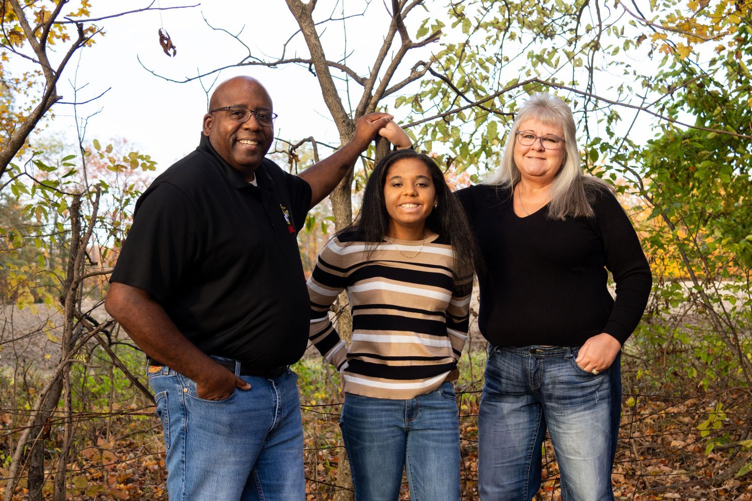 Family of three posing together in a wooded autumn setting with bare branches and colorful fall leaves surrounding them.