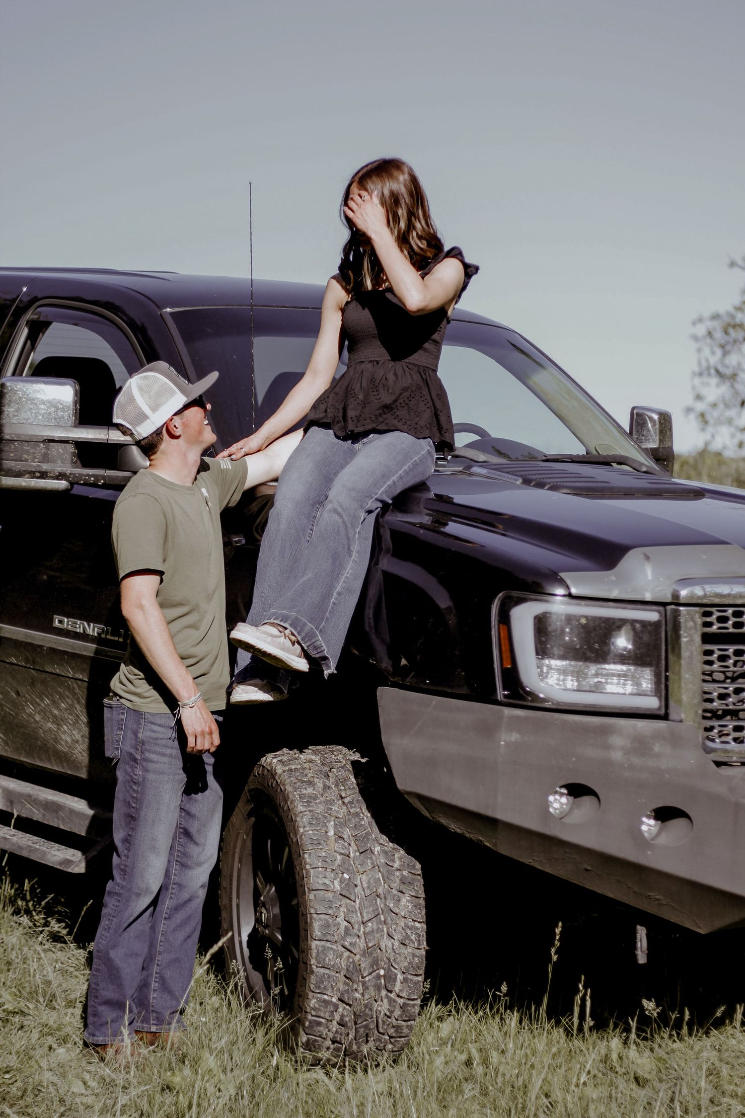 A young man in a cowboy hat helps a girl sitting on the hood of a lifted black pickup truck.