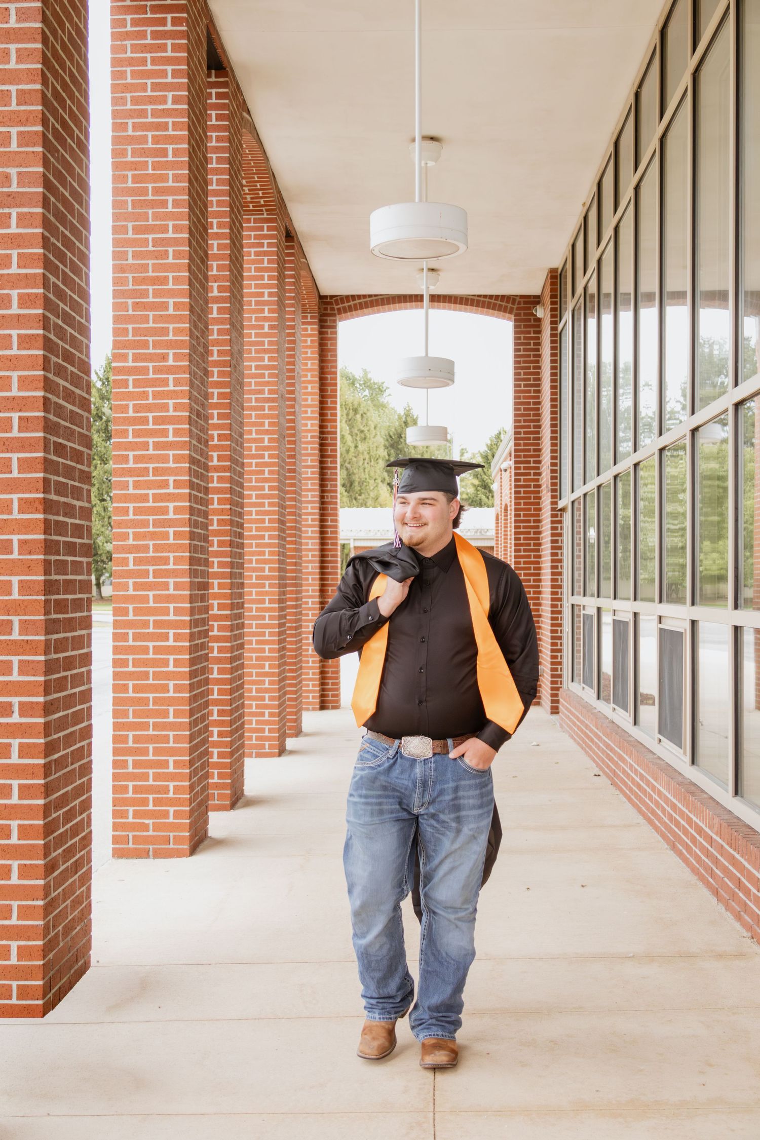 Smiling graduate in cap and gown walking through brick school corridor carrying graduation robe.