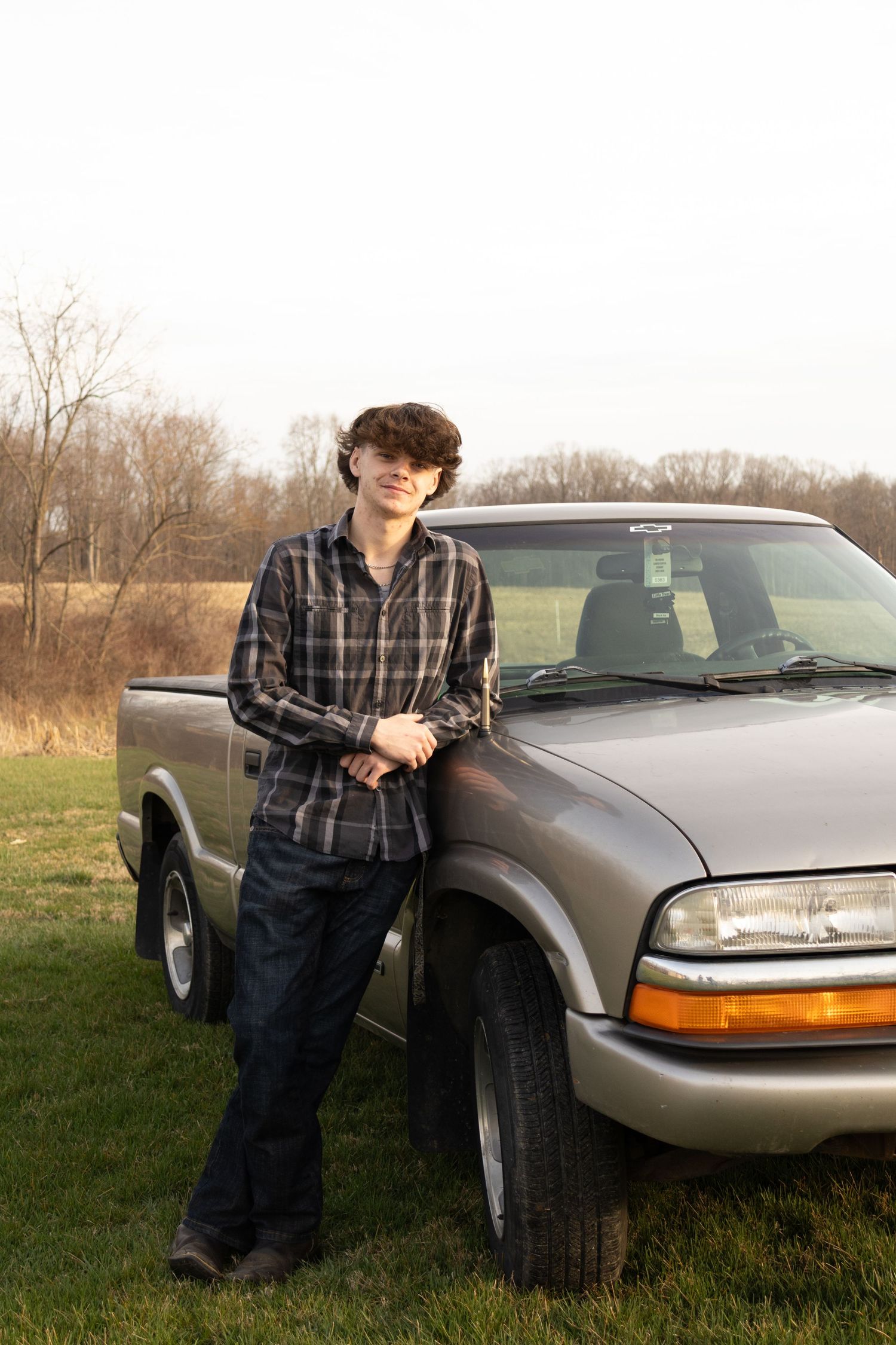 Teen boy in plaid shirt leaning against silver Chevy S10 pickup truck in grassy field.