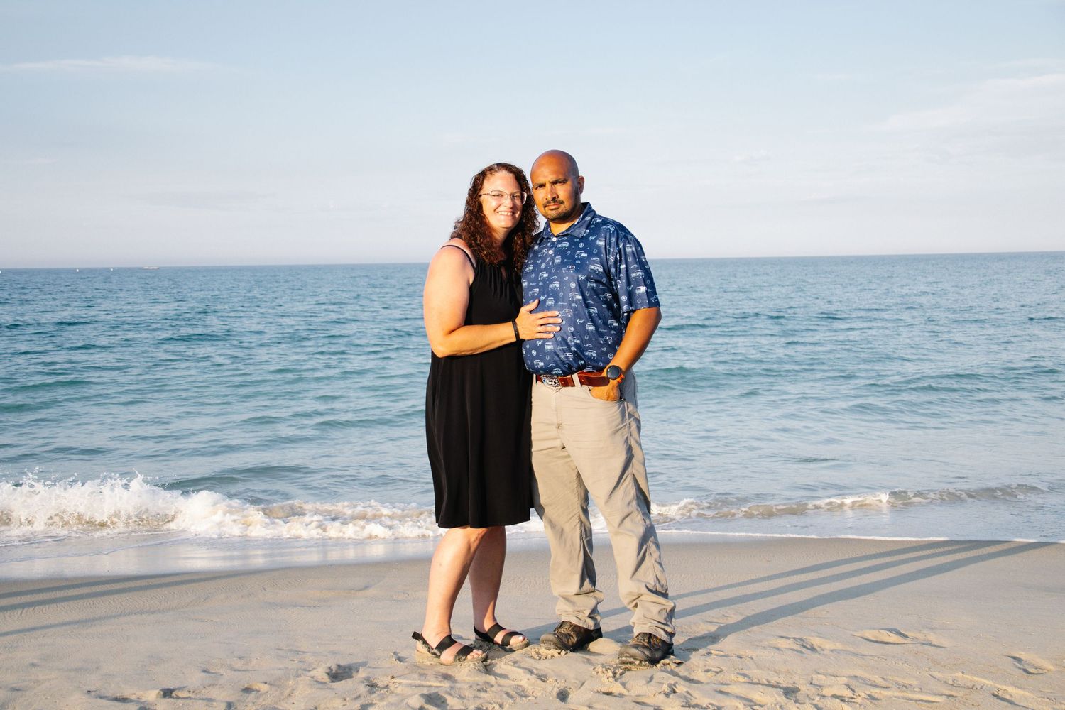 Smiling couple embracing on a sunny beach with calm ocean waves behind them.