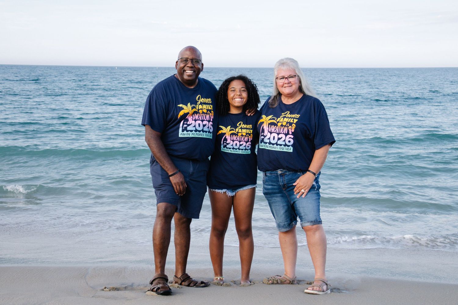 Three family members stand together on a sandy beach wearing matching navy blue Jones Family Vacation 2026 t-shirts.