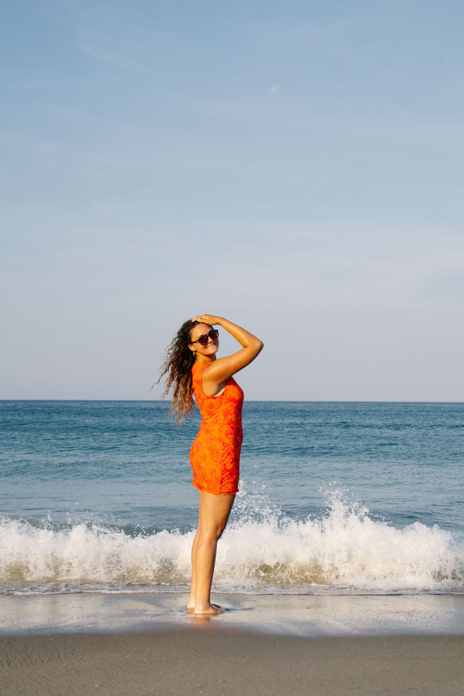 Woman in orange dress shielding eyes from sun while standing in ocean waves on a sunny beach.